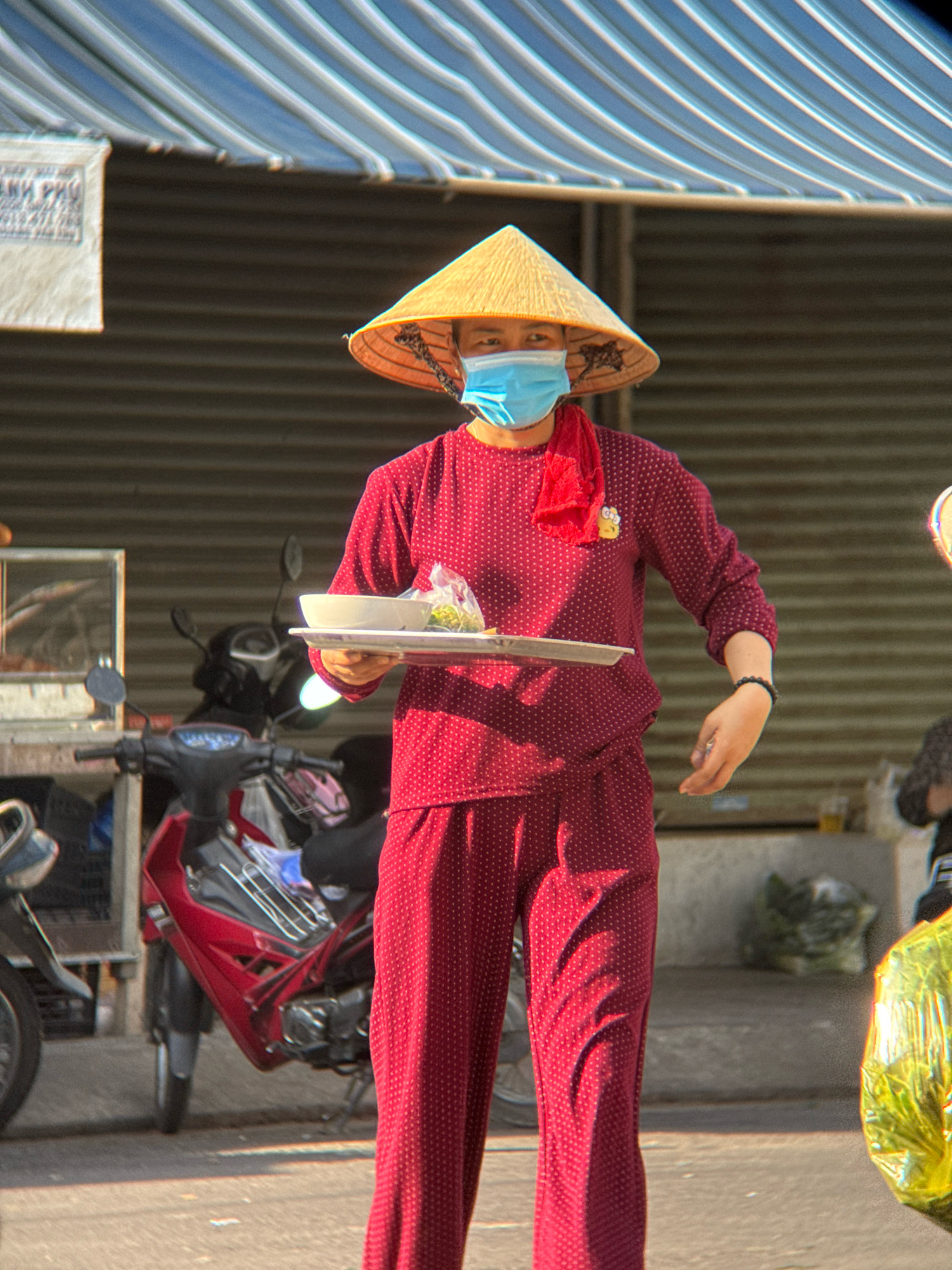 In Nha Trang, Vietnam, a person wearing a conical hat and a blue face mask is walking. They are dressed in a red polka-dot shirt and matching pants. In their hands, they hold a white bowl and a plastic bag of food. The scene is set outdoors, with a metal shuttered building in the background and motorcycles parked nearby. A striped awning is visible overhead.