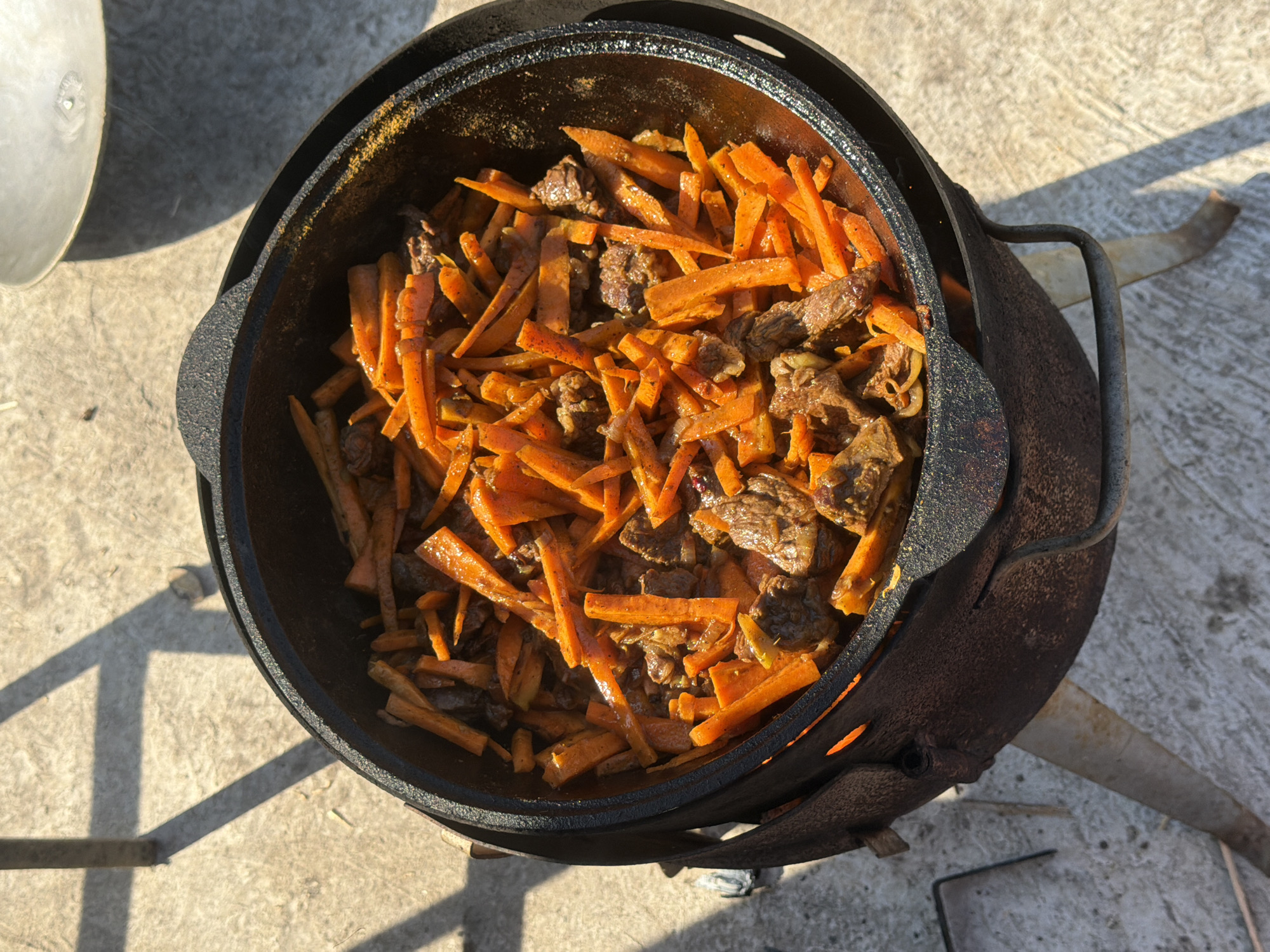 The image, likely taken outdoors in daylight in Большая Шильна, Russia, shows a cast iron pot, presumably a kazan, containing a dish of meat and julienned carrots. The pot is positioned on a stove or stand of similar material. The ground is concrete. A partial view of a lid sits to the left. The scene is still, with no apparent movement.