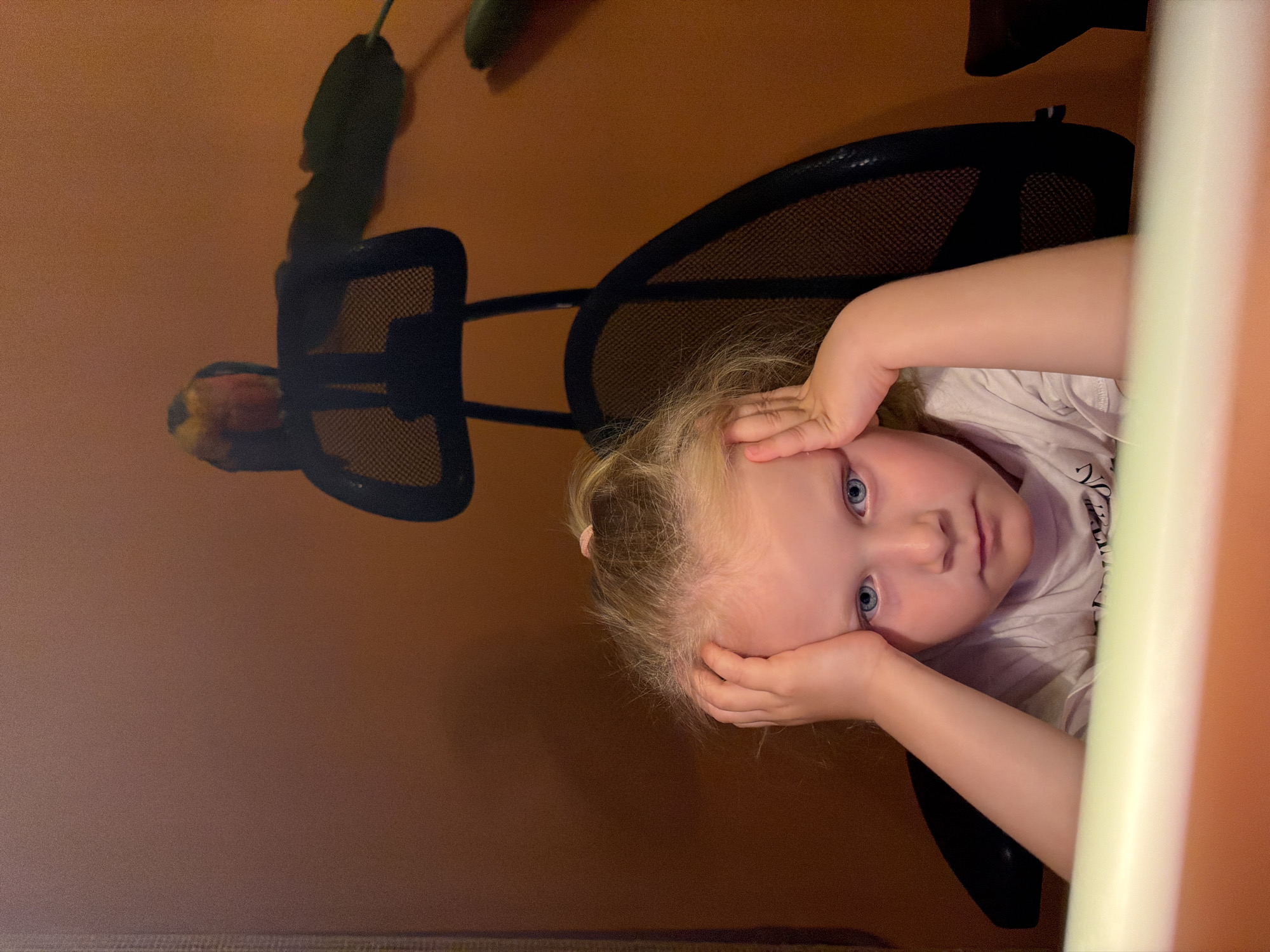 A young, blonde-haired child with blue eyes is seated in a black mesh chair. She is holding her hands to her forehead. The background includes a brown wall and a plant. The photo appears to be taken from an unusual angle. There is no verifiable information to indicate that the photo was taken in the city of Khimki, Russia.