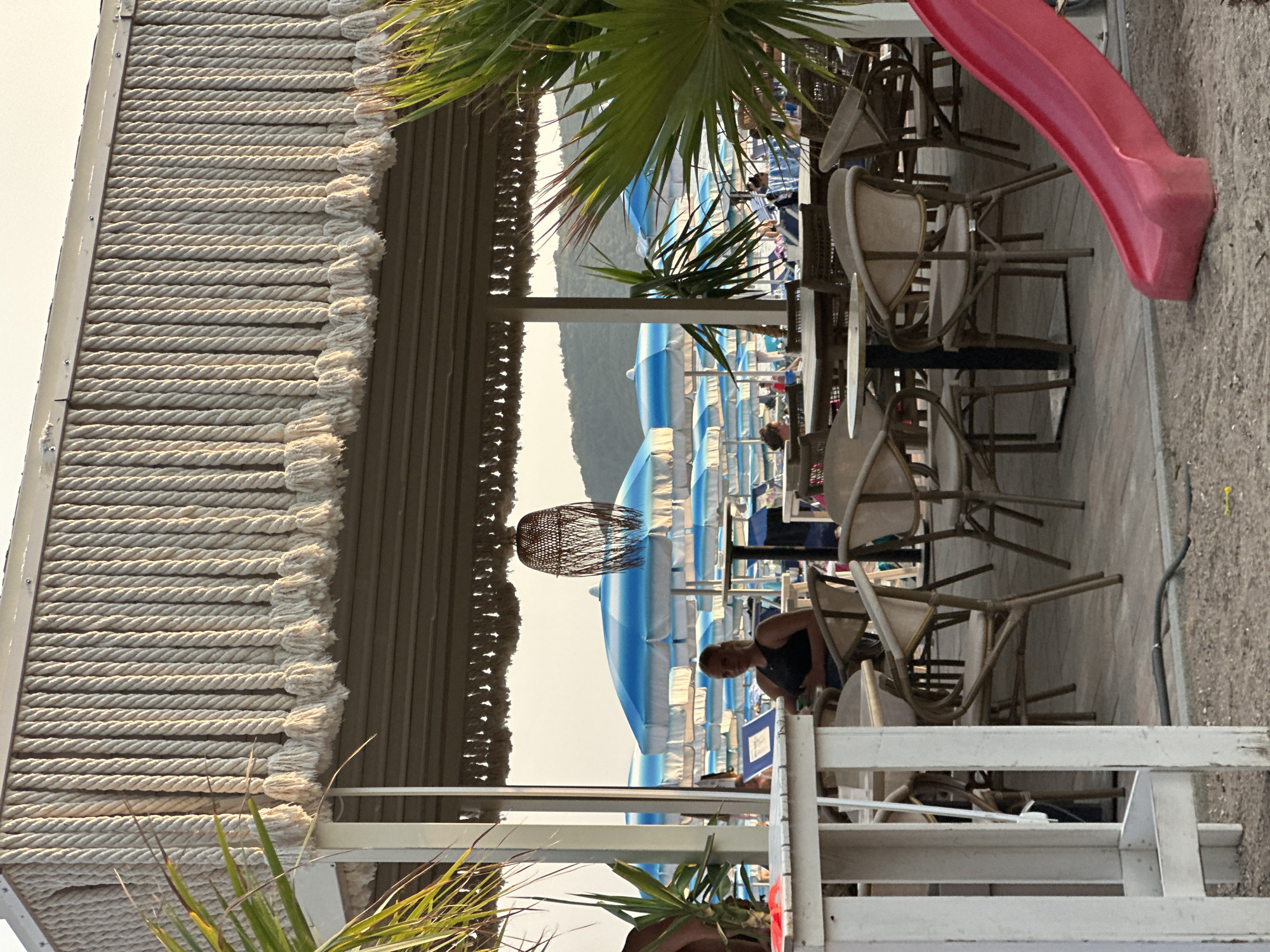 The image depicts a beachside cafe in Boreti, Montenegro. In the foreground, a white wooden table and chairs are visible, with a red slide positioned to the right. The cafe's roof is adorned with rope detailing. Beyond the cafe, rows of blue and white umbrellas line the beach, with several people seated at tables and scattered around. The background features green hills under a hazy sky. A woman wearing a black tank top is seated at one of the cafe tables.