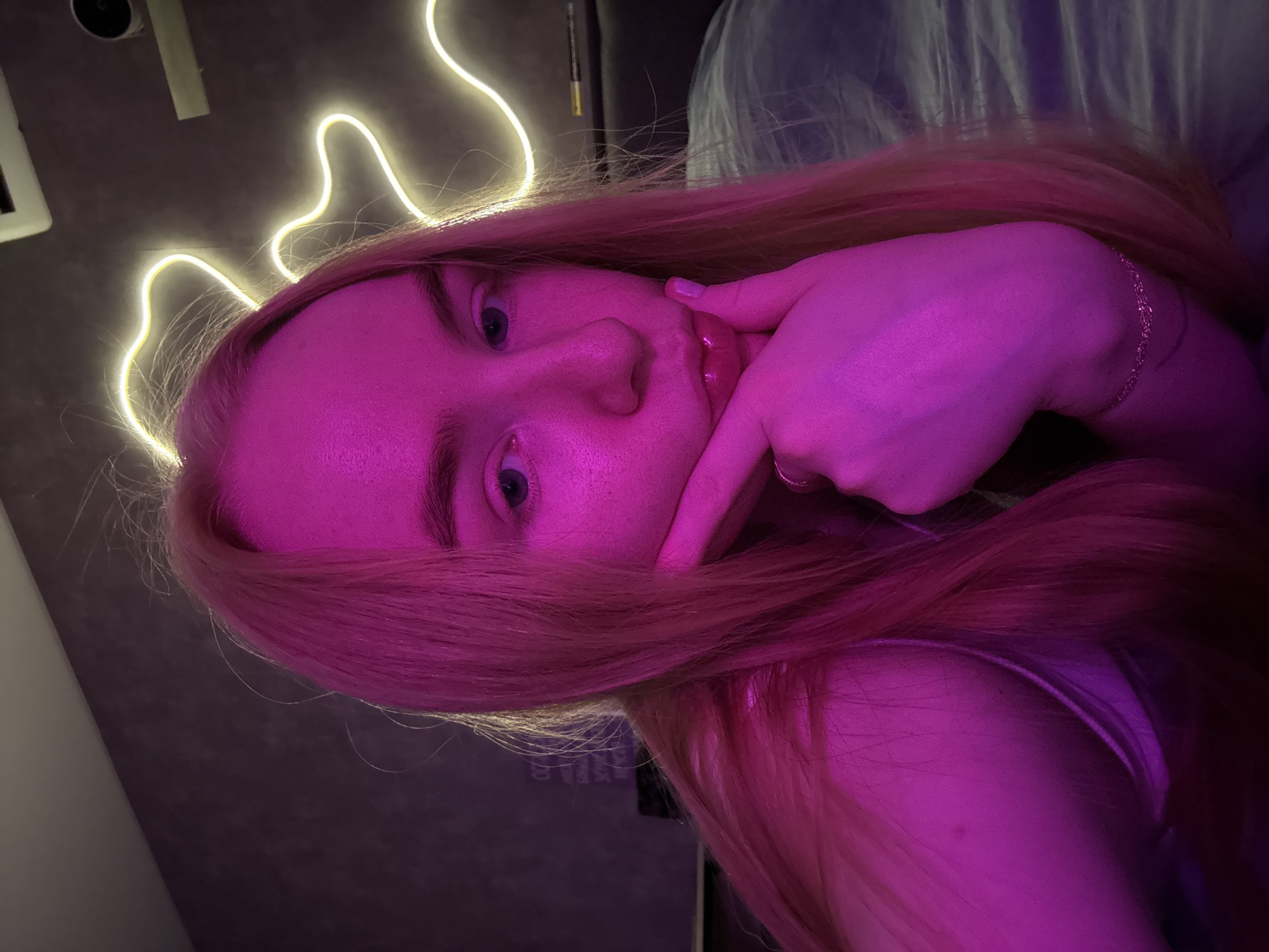 A young fair-skinned individual with light hair poses indoors with a neon light above their head. The individual's face is partly illuminated by pink and purple lighting. The person's left hand is touching their face. A piece of furniture or bedding is located in the background. This image could be from Chaikovsky, Russia, but this is not discernible from the visual data.