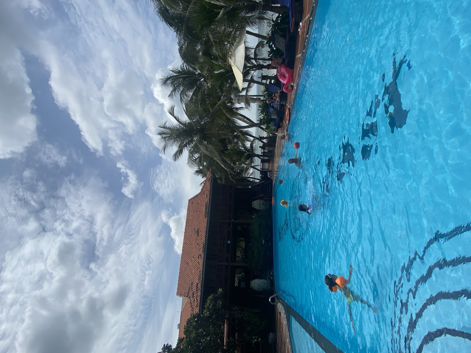 The image depicts a resort swimming pool in Mũi Né, Vietnam, on a partly cloudy day. The pool is filled with clear blue water, and its edges are lined with a tiled walkway. Several people are visible in the pool: a young girl with orange arm floaties is swimming, and other individuals, including children and adults, are present. Some are swimming, while others appear to be relaxing in the water.
Around the pool, there are lounge chairs, some occupied by people. A pink inflatable ring shaped like a flamingo is visible near the pool's edge, along with a white umbrella providing shade. Lush green palm trees frame the scene, with the ocean visible in the background. Behind the pool area, a large building with a red-tiled roof and wooden accents stands, suggesting it is part of the resort's accommodation or dining facilities. The sky is a mix of blue with scattered white clouds.