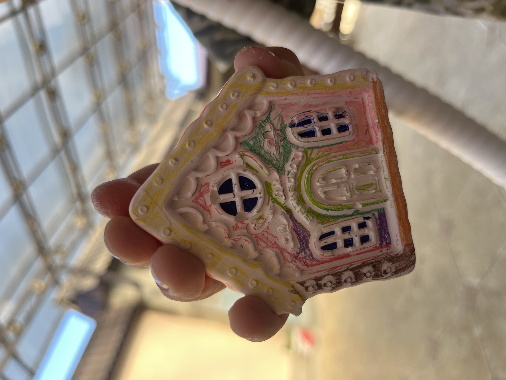 A person is holding a small, decorated house-shaped object. The house is predominantly white with colorful details including pink, green, yellow, purple, orange, and brown. It features designs mimicking windows and trim. The person's fingers are visible holding the object. The background includes a blurry view of what appears to be a building's window structure and a light-colored surface. The city of Kaspiysk, Russia is not recognizable in the image.