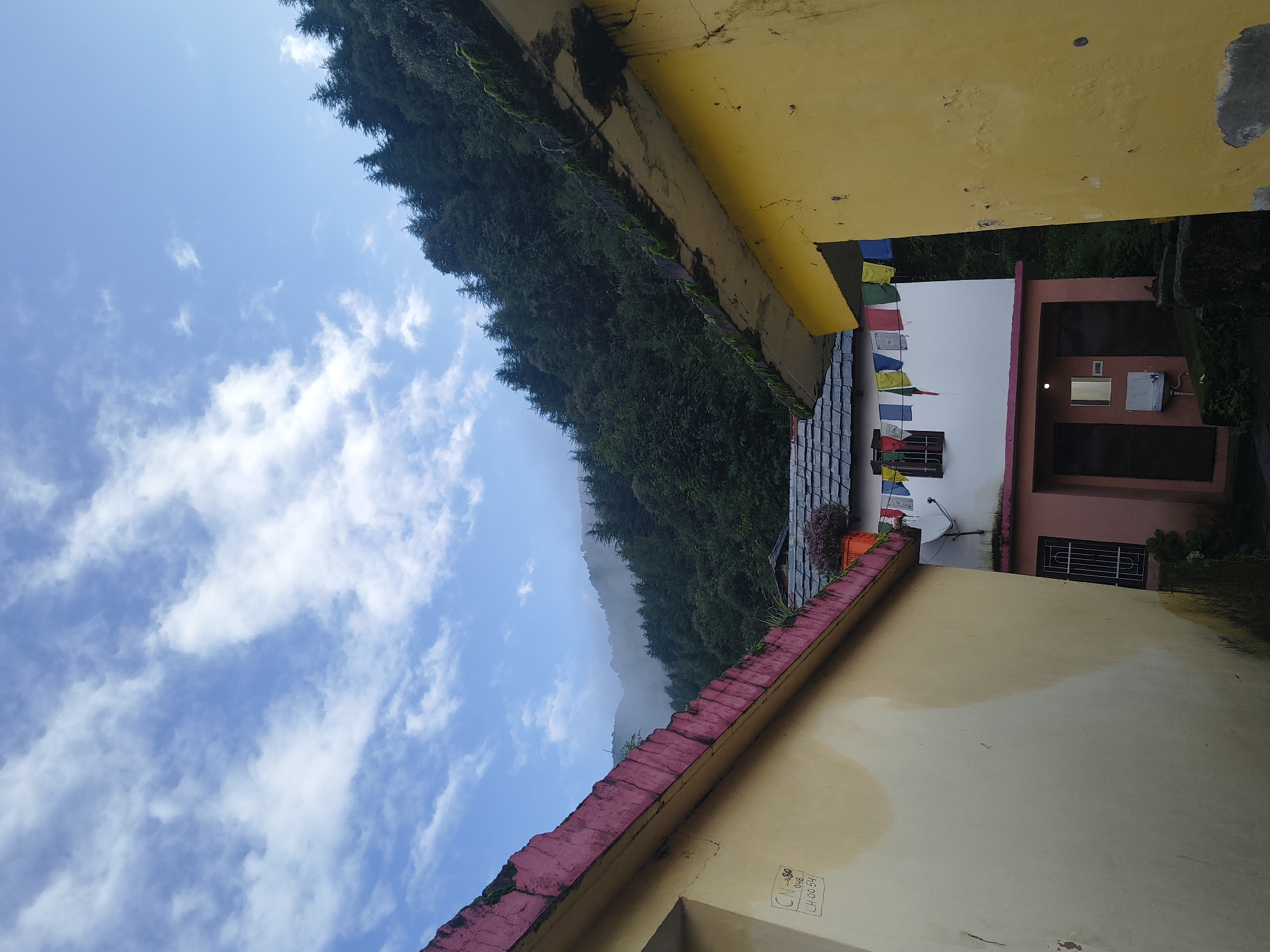 The image captures a serene view in the city of Haini, India, characterized by a tranquil mountain landscape. In the foreground, two buildings are partially visible, their walls painted in pale yellow and pink hues. The roofs of these structures are adorned with prayer flags, adding a touch of vibrant color to the scene. Beyond the buildings, a lush, green forest blankets the mountainside, with a gentle mist clinging to the higher elevations. The sky above is a brilliant blue, dotted with scattered white clouds, creating a picturesque backdrop. The overall atmosphere is one of peace and natural beauty.