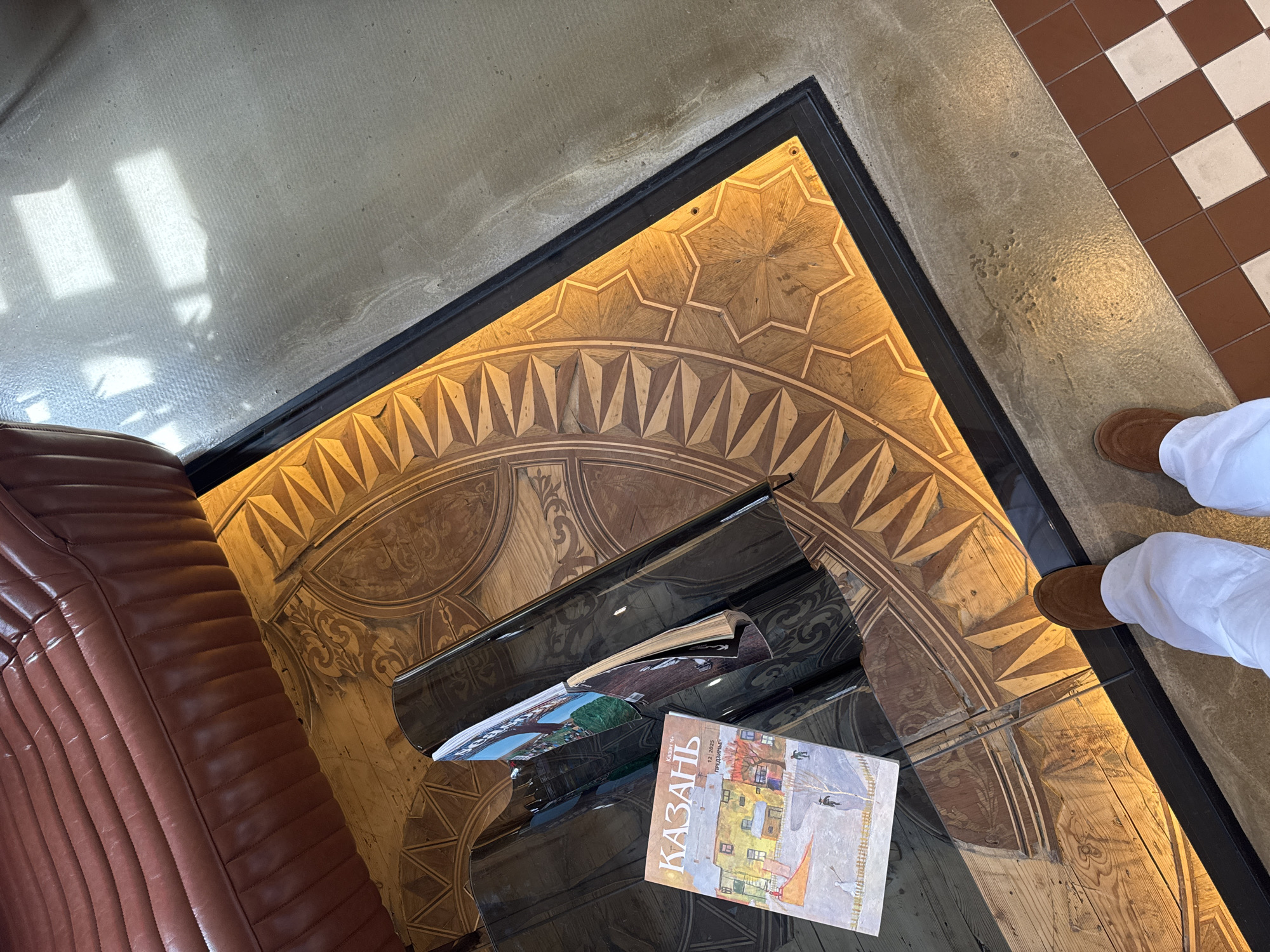 The image shows an interior in Kazan, Russia. A brown leather sofa is visible on the left. In the center, a black glossy table displays magazines and books, including one with the word "Казань" (Kazan) on the cover. The floor has intricate wooden patterns and designs, framed by a black border. To the right, a person wearing brown shoes and white pants stands partially visible. The background features a mix of flooring materials, including a beige, textured surface and a brown and white checkered pattern.