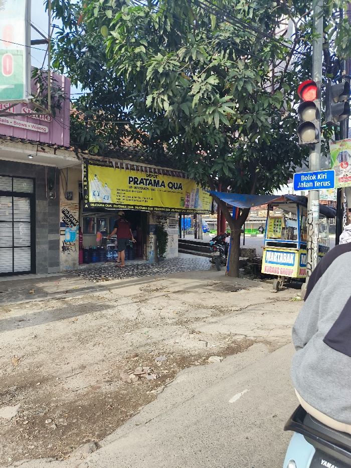 The image shows a street scene in Soreang, Indonesia, located at the coordinates -7.02 latitude and 107.541 longitude.  A man wearing a blue shirt walks in front of a store with a yellow sign that reads "DEPOT PRATAMA QUA."  A traffic light with a red light is visible. There is a sign in Indonesian that reads "Belok Kiri Jalan Terus" which means "Turn Left, Go Straight." A small street vendor cart is parked on the street with a sign that reads "MARTABAK KUDANG GROUP." A motorcycle is parked in the foreground. The road is made of asphalt and appears to be in need of repair. The street is bordered on one side by a building with a pink facade. A large tree grows in the foreground.  The image is taken from a low angle and the composition is centered on the street.  The image is well lit, with the sun shining brightly.  The scene is vibrant and active.