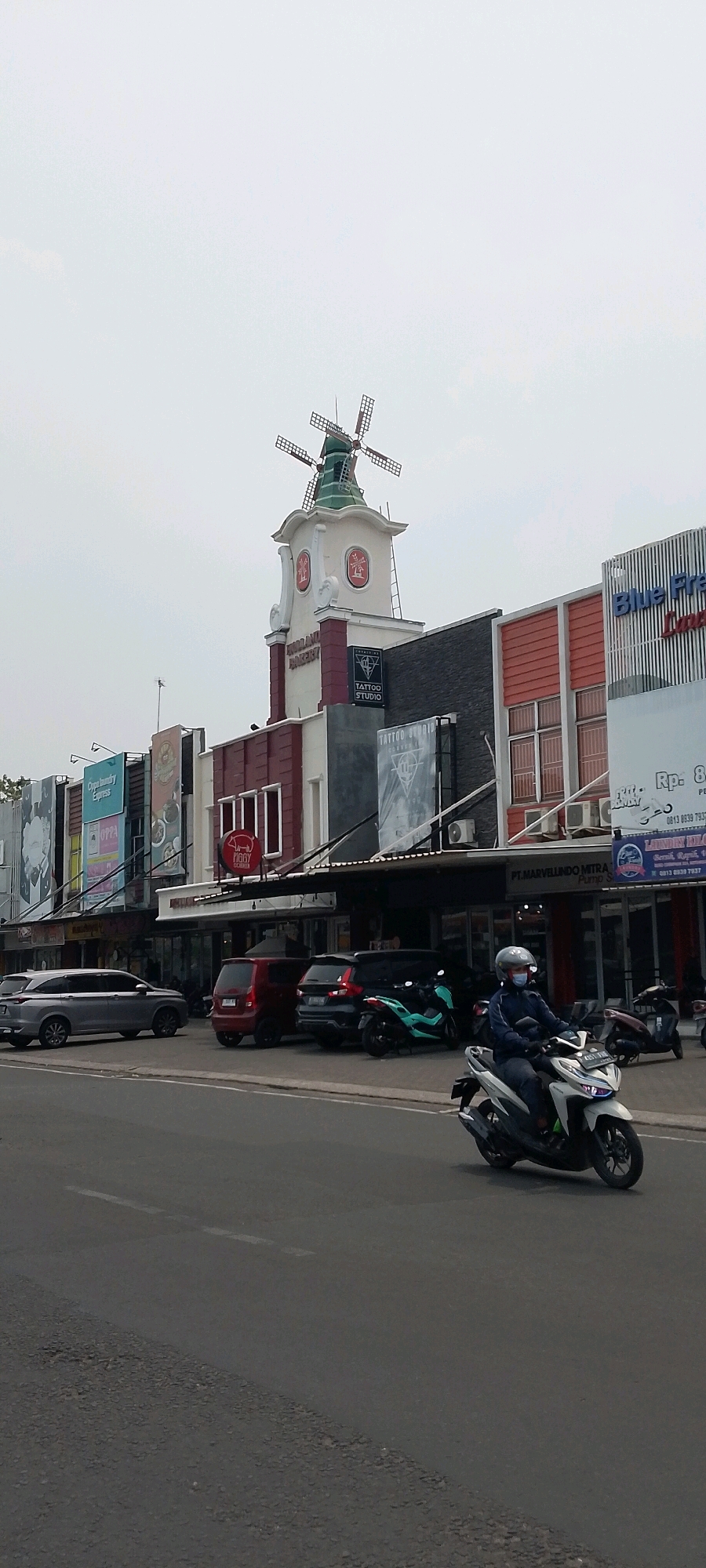 In Cileungsi, Indonesia, a street scene unfolds with a prominent building featuring a Dutch-style windmill atop its tower. The building is a commercial establishment with various businesses visible, including a "Tattoo Studio" and "Blue Fresh Laundry". Several vehicles are parked in front of the shops: a silver SUV, a red hatchback, a dark SUV, and a teal scooter. A person wearing a helmet and mask rides a white scooter in the foreground, moving from right to left across the frame. The sky is overcast, and the road surface shows some wear and tear.