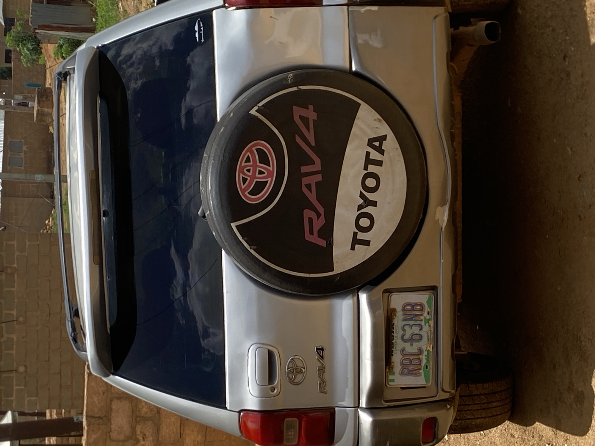 A silver Toyota RAV4 SUV is parked in what appears to be a residential area in Bukuru, Nigeria. The rear of the vehicle is visible, showing the spare tire cover with the Toyota and RAV4 logos. The license plate, reading "RBC-63NB," is attached to the vehicle's rear. Buildings constructed of brown brick can be seen in the background, along with some vegetation. The ground surrounding the car is dirt or sand.