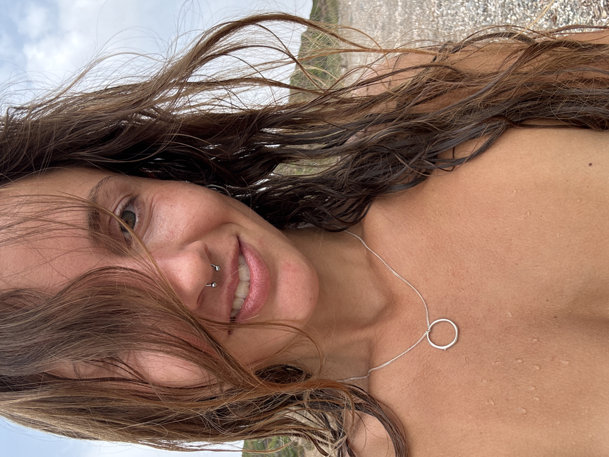 A close-up, over-the-shoulder selfie of a young woman with long, wet, brown hair is taken outdoors on a beach. She has a septum piercing and is wearing a silver necklace with a circular pendant. Her skin is tanned and appears wet. In the background, the sky is cloudy and there is a glimpse of a sandy, pebbly beach. The woman is smiling.