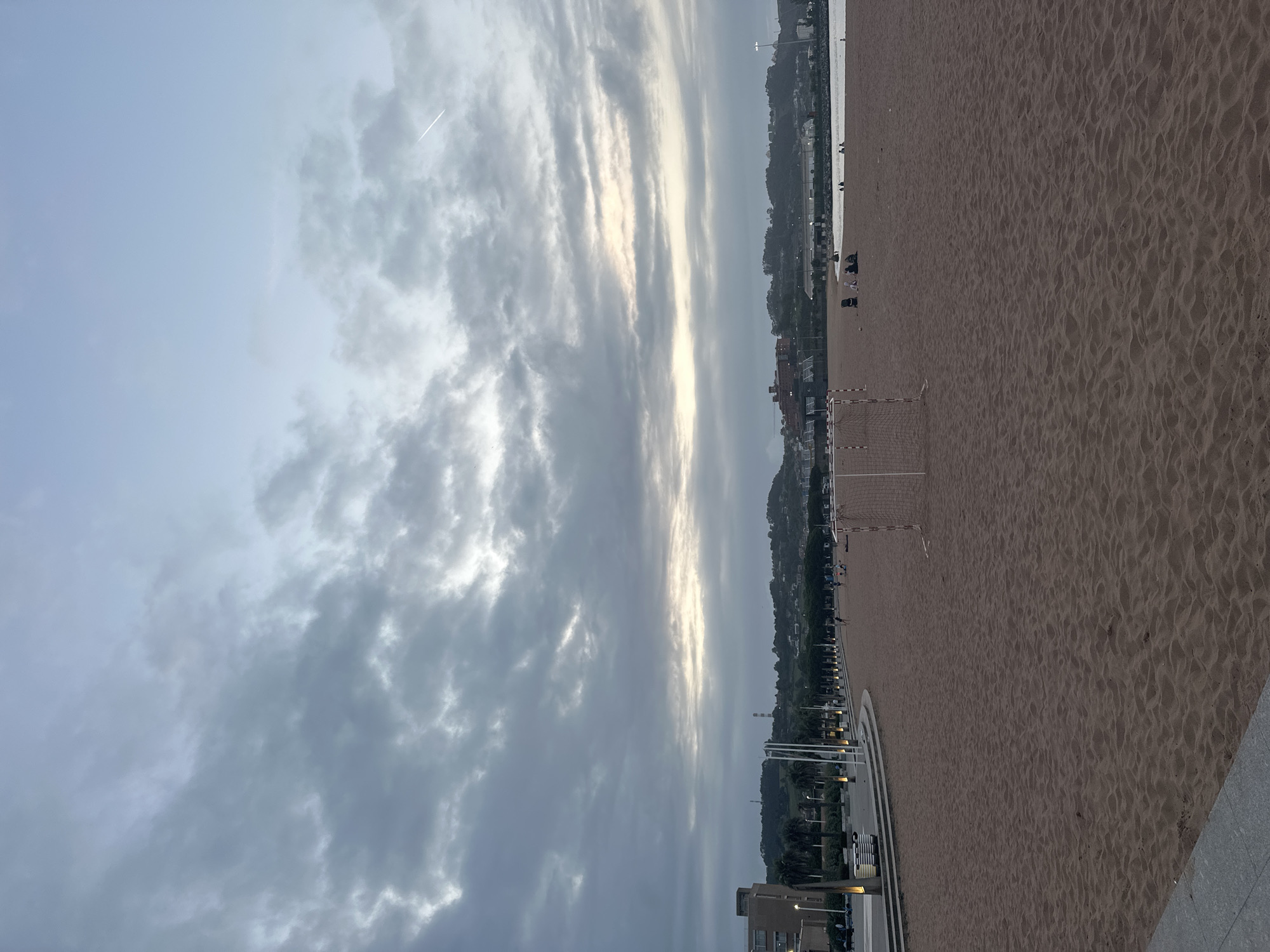 The image presents a wide view of San Lorenzo Beach in Gijón/Xixón, Spain, during dusk or dawn under an overcast sky. The foreground is dominated by a vast expanse of reddish-brown sand exhibiting subtle undulations. In the mid-left area of the beach, a white-framed portable soccer goal with a net stands on the sand. Further along the beach towards the right, multiple small groups of individuals are visible; one group of approximately three to four people is seated on the sand near the water's edge, adjacent to a dark, low object. A calm, dark blue-grey body of water (the sea) borders the beach on the right side.
A paved promenade runs parallel to the beach on the left, separated by a multi-tiered concrete structure. This promenade is lined with regularly spaced modern light poles emitting a warm glow, with some stacked white loungers positioned near the poles. Behind the promenade, a multi-story building with a light-colored facade is visible, partially obscured by palm trees. The background features a series of green, undulating hills covered in vegetation, interspersed with various urban buildings. Distant industrial structures or towers are faintly visible against the horizon line on the hills. The sky is predominantly covered by grey, textured clouds, with a brighter band of light visible near the horizon, suggesting the position of the sun. Patches of pale blue sky are discernible in the upper regions, and a faint white airplane contrail streaks across the upper right quadrant. Artificial lights from the promenade and distant structures are active, indicating low ambient light conditions.