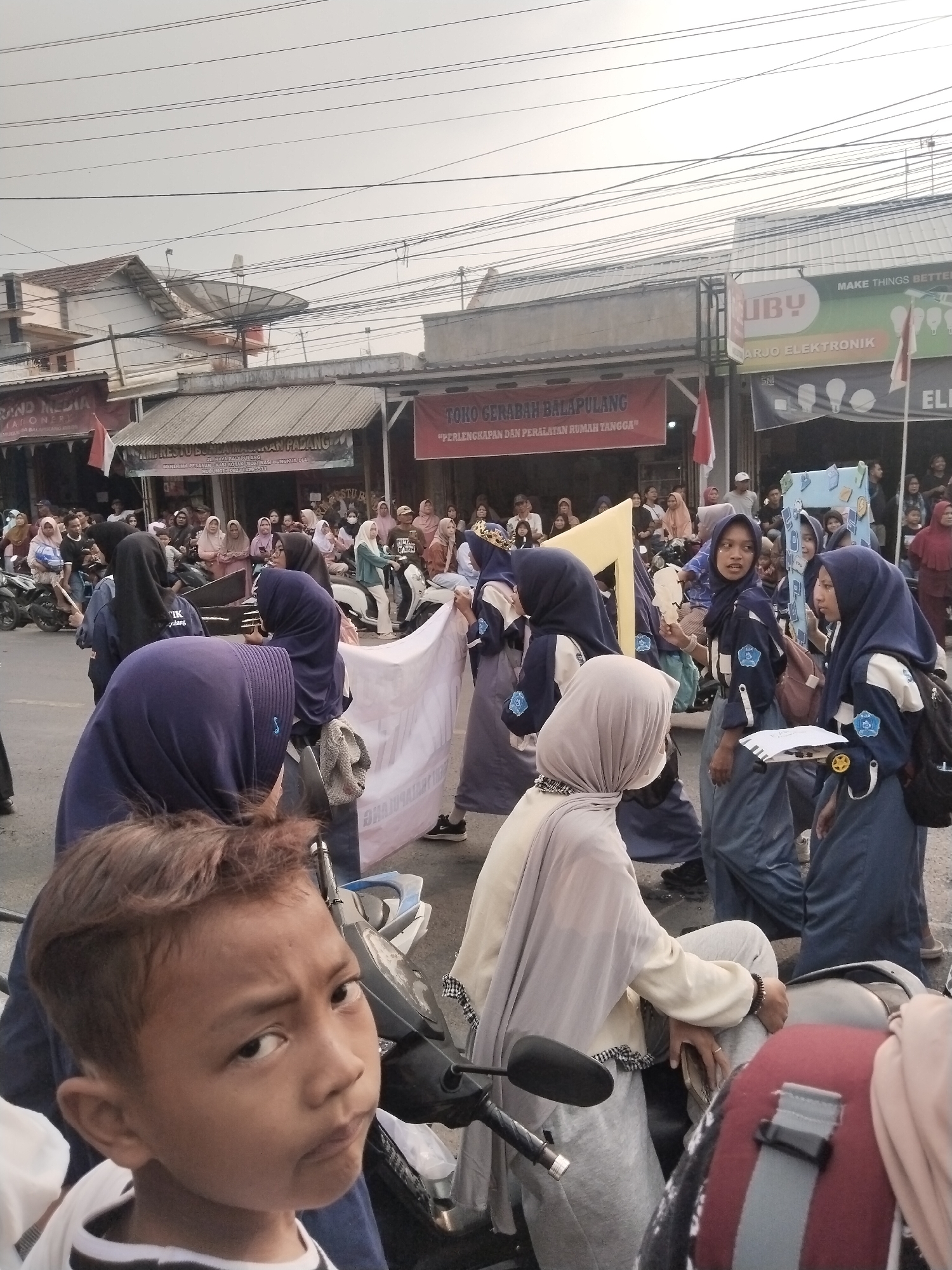 The image depicts a street scene in Balapulang Kulon, Indonesia, likely during a procession or event. In the foreground, a young boy with brown hair is looking directly at the camera with a neutral expression. He is wearing a black and white striped shirt. Behind him, several individuals in school uniforms, primarily female students wearing hijabs and blue and white uniforms, are present. Some are holding a large white banner with text on it, while others are carrying props or walking. The background reveals a street lined with buildings, including shops with signage such as "UBY NARJO ELEKTRONIK" and "TOKO GERABAH BALAPULANG." There are several motorcycles parked along the street, and a crowd of onlookers is visible further down. Overhead, numerous electrical wires crisscross the sky. The overall atmosphere suggests an outdoor public gathering.