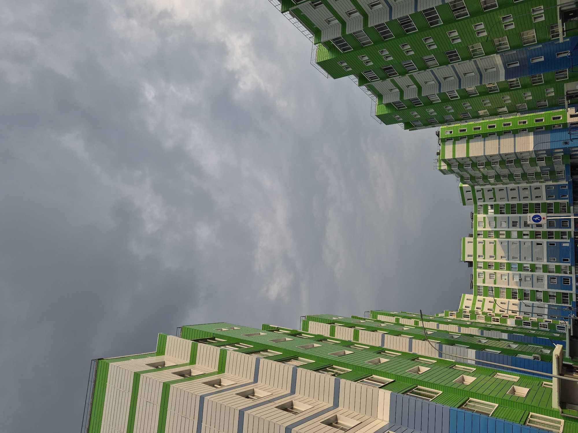The image shows a low-angle shot of modern apartment buildings in Nyagan, Russia, against a cloudy sky. The buildings are tall and feature a distinctive architectural style with repeating geometric patterns in green, white, and blue. The facades are clad in panels, with numerous windows visible. The perspective is from below, looking upwards towards the sky, creating a sense of scale and vastness. The sky is filled with heavy, gray clouds, suggesting overcast weather, possibly before or after rain. The lighting is diffused, with some areas of the buildings catching subtle sunlight, hinting at a time around late afternoon or early morning. No people are visible in the image. There are no specific event cues, and the scene is static. One visible sign has Cyrillic text and an international symbol. The overall impression is one of contemporary urban architecture set against a dramatic, moody sky.
