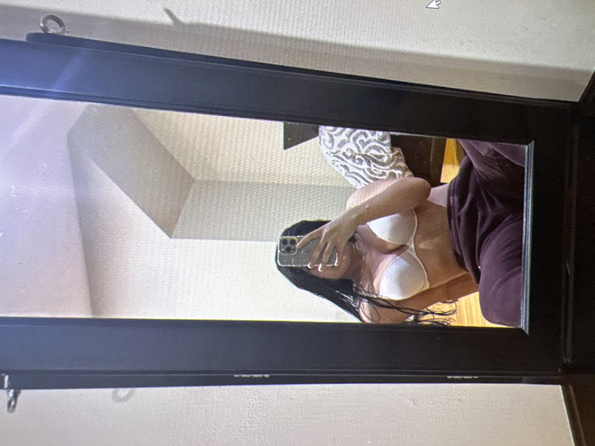 The image shows a person taking a mirror selfie. The person is wearing a white bra and dark purple sweatpants. They are sitting on the floor in front of a large, dark-framed mirror. The mirror reflects the person, a bed with a patterned duvet, and a portion of the room. The room appears to be a bedroom. The image is not geo-tagged and provides no information about the location in Castelo Branco, Portugal.