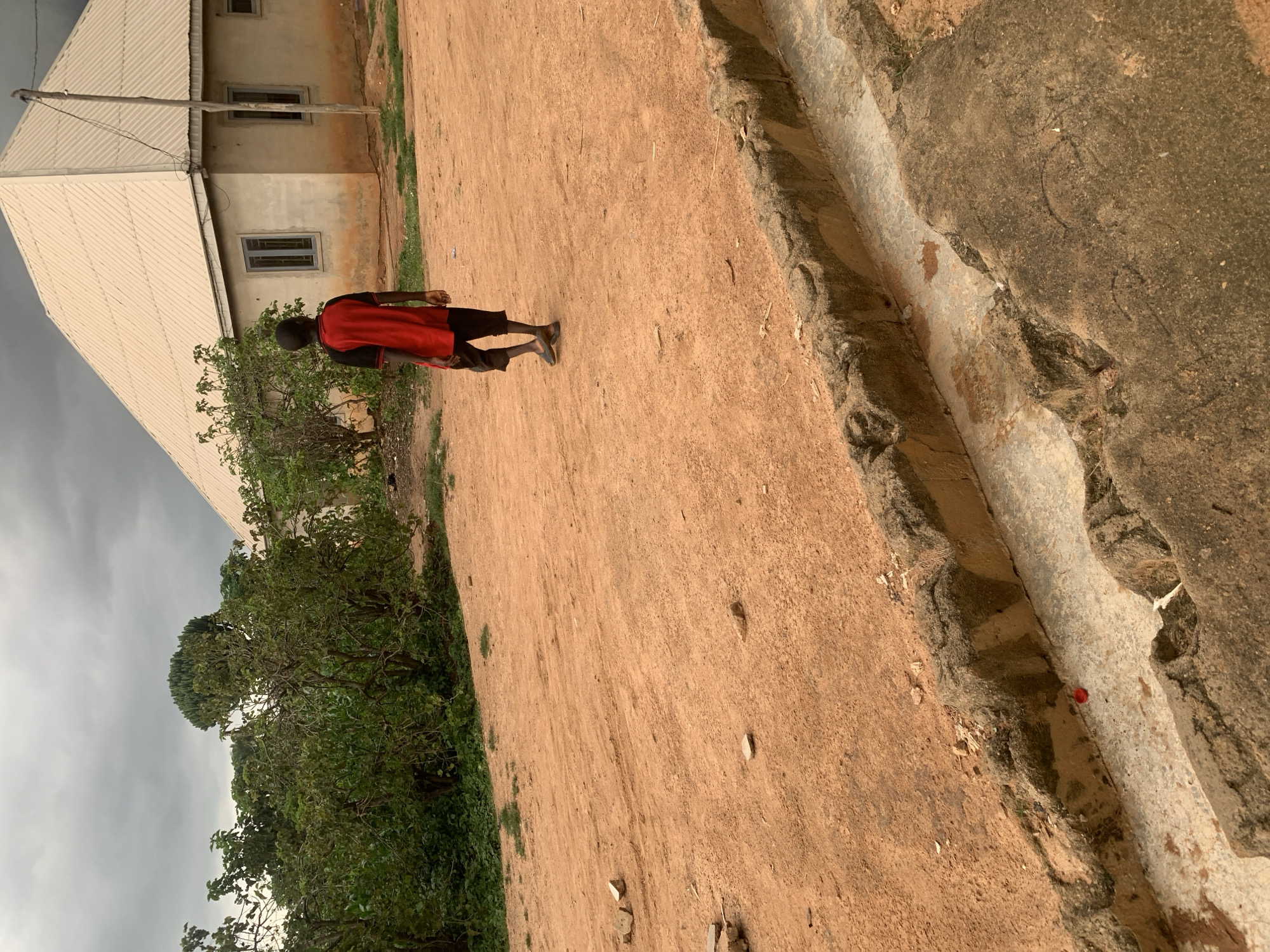 A young boy wearing a red and black t-shirt and dark shorts walks away from the camera on a reddish-brown dirt path in Sabon Tasha, Nigeria. He is heading towards a pale yellow, square building with a light beige, corrugated metal roof. Two dark-framed windows are visible on the side of the building facing the boy. Green bushes and trees are to the left of the boy and the building. A grey, overcast sky hangs above the scene. In the foreground, a concrete drainage ditch runs horizontally across the image, with rough concrete edges. A small, red object, possibly a bottle cap, lies in the ditch.