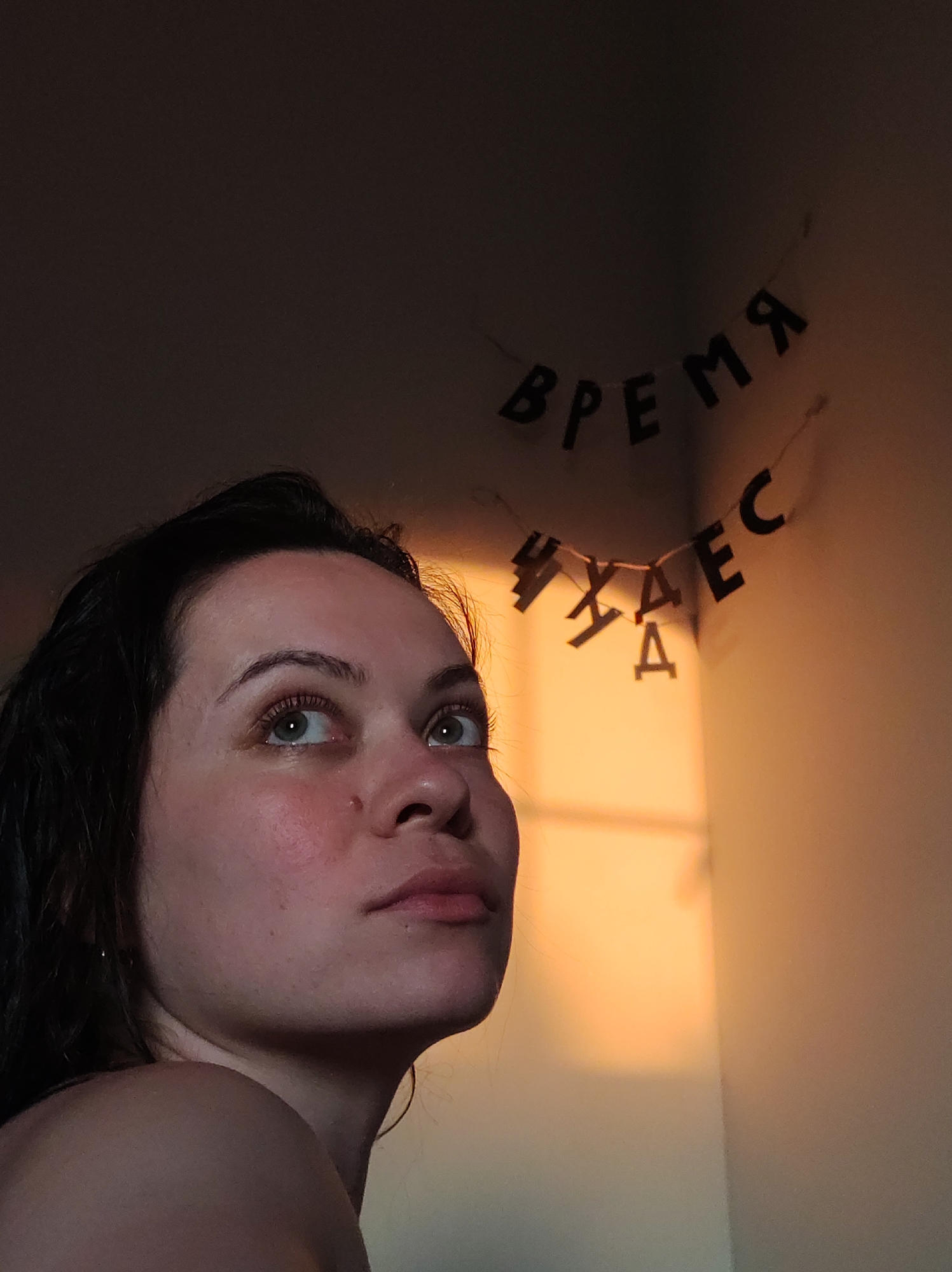 A low-angle shot shows a woman with wet, dark hair looking upward towards a decorative string of letters. The letters, which appear to be made of black cardboard, read "ВРЕМЯ ЧУДЕС" (Time of Miracles) in Russian. The woman has fair skin, green eyes, and a slight flush on her cheek. She is wearing minimal clothing, exposing her shoulder. The background is a plain wall with a gradient of light, transitioning from dark in the upper left corner to a warm, orange glow in the lower right corner. The location is indoors, but the city is unknown.