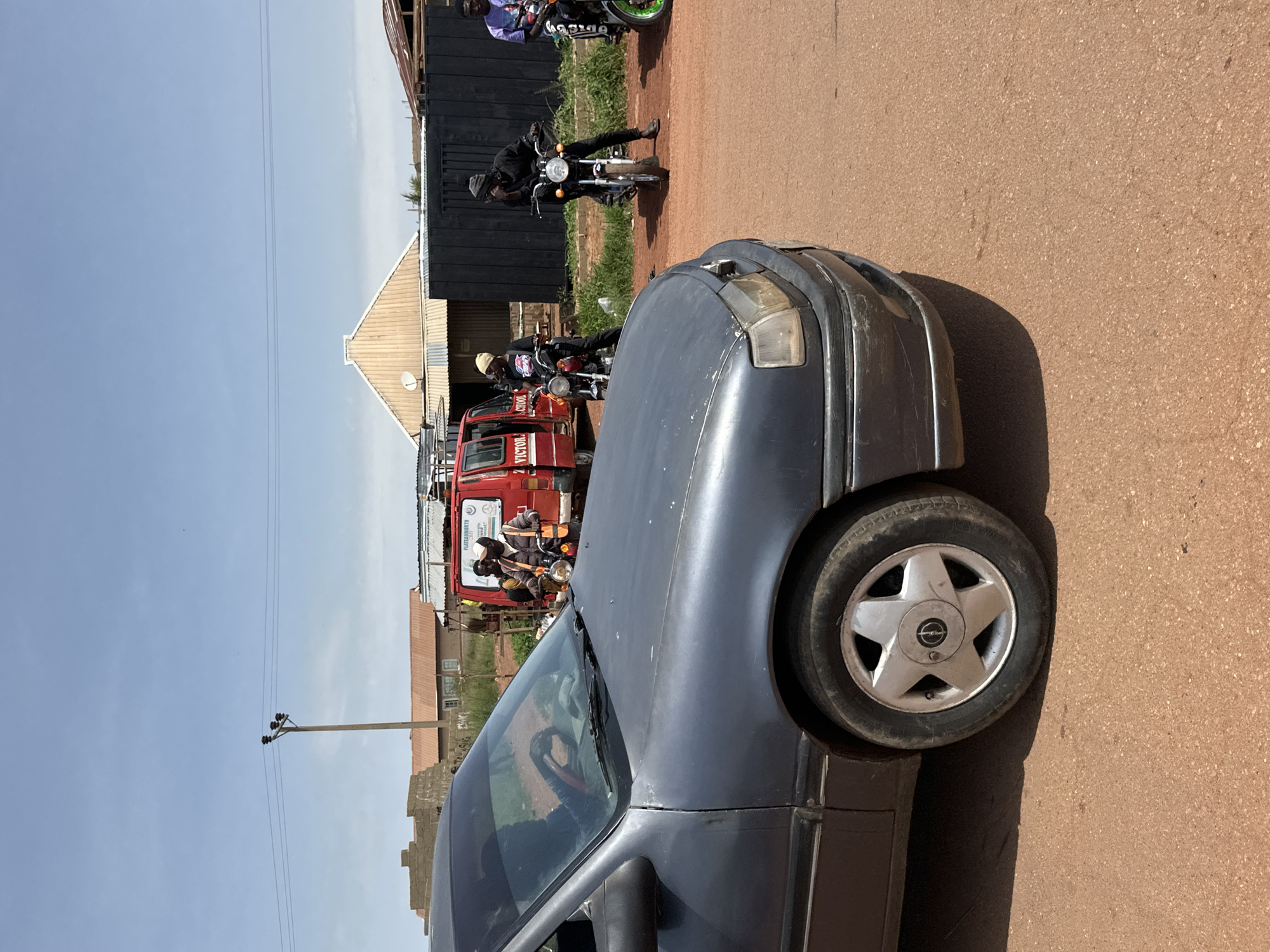 A dark grey sedan is parked on a dirt road, partially obscuring a scene with a red van and several individuals on motorcycles. The sedan's front left wheel and bumper are visible. The red van, labeled "VICTOR SCHOOL," is stopped, and a person wearing a reflective vest is standing next to it, looking forward. Three men are seated on motorcycles behind the van. The man on the leftmost motorcycle is wearing a black beanie and a black jacket. The man in the middle motorcycle is wearing a black beanie and a black outfit. The man on the rightmost motorcycle is wearing a purple shirt. The environment appears to be an urban setting in Nigeria, characterized by dusty roads, modest buildings in the background, and power lines overhead. The sky is clear and blue.