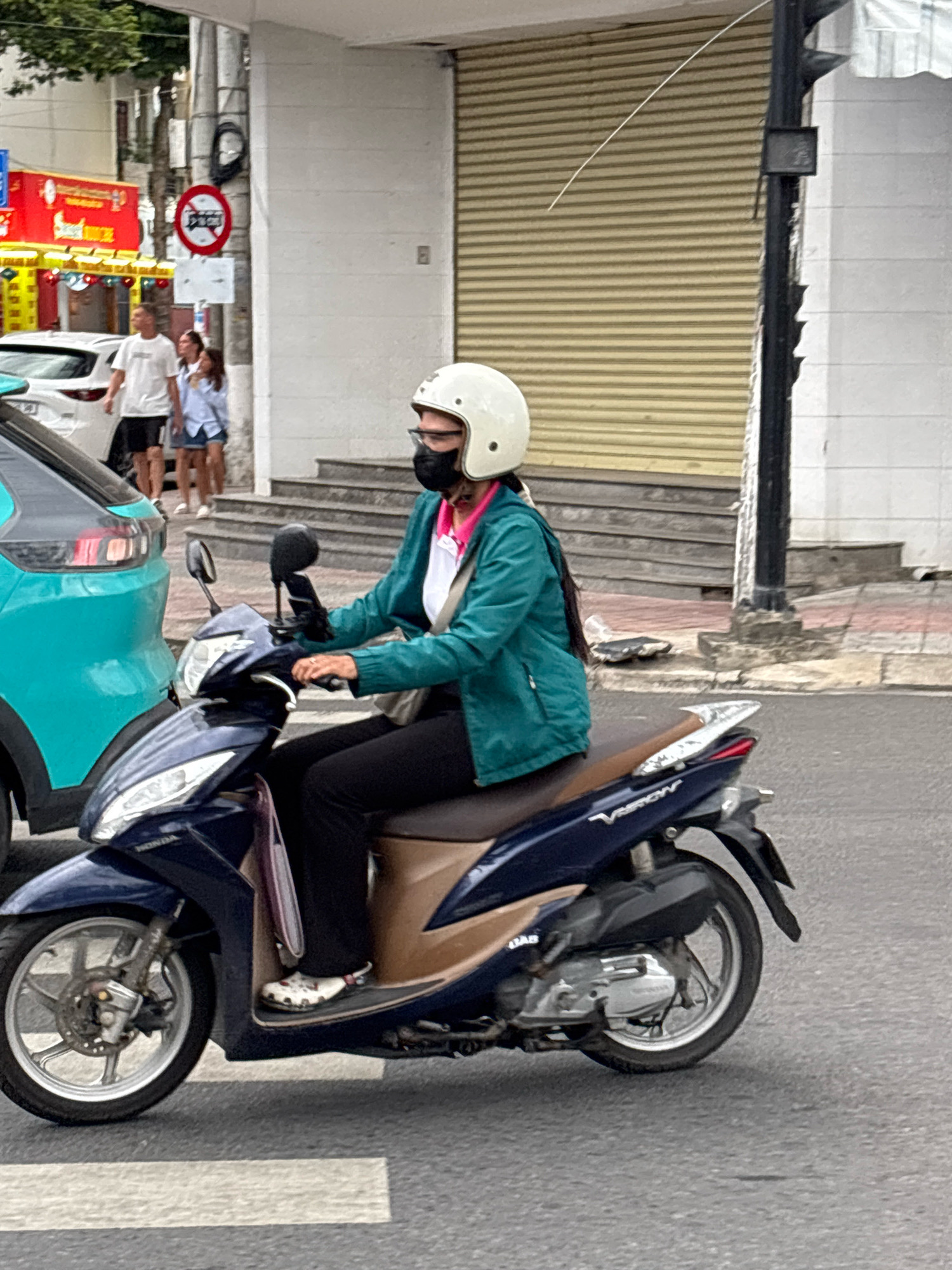 A person wearing a white helmet and a teal jacket rides a dark blue scooter on a street in Nha Trang, Vietnam. The person is wearing a black mask and a pink garment around their neck. A light-colored bag is slung across their body. The scooter has a brown seat and silver accents. In the background, there are buildings with beige roll-up doors, a street light pole, and a white car. To the left, a turquoise vehicle is partially visible. A red sign with a white circle and a diagonal line, indicating a prohibition, is mounted on a pole. A pedestrian crossing is marked on the asphalt in the foreground.