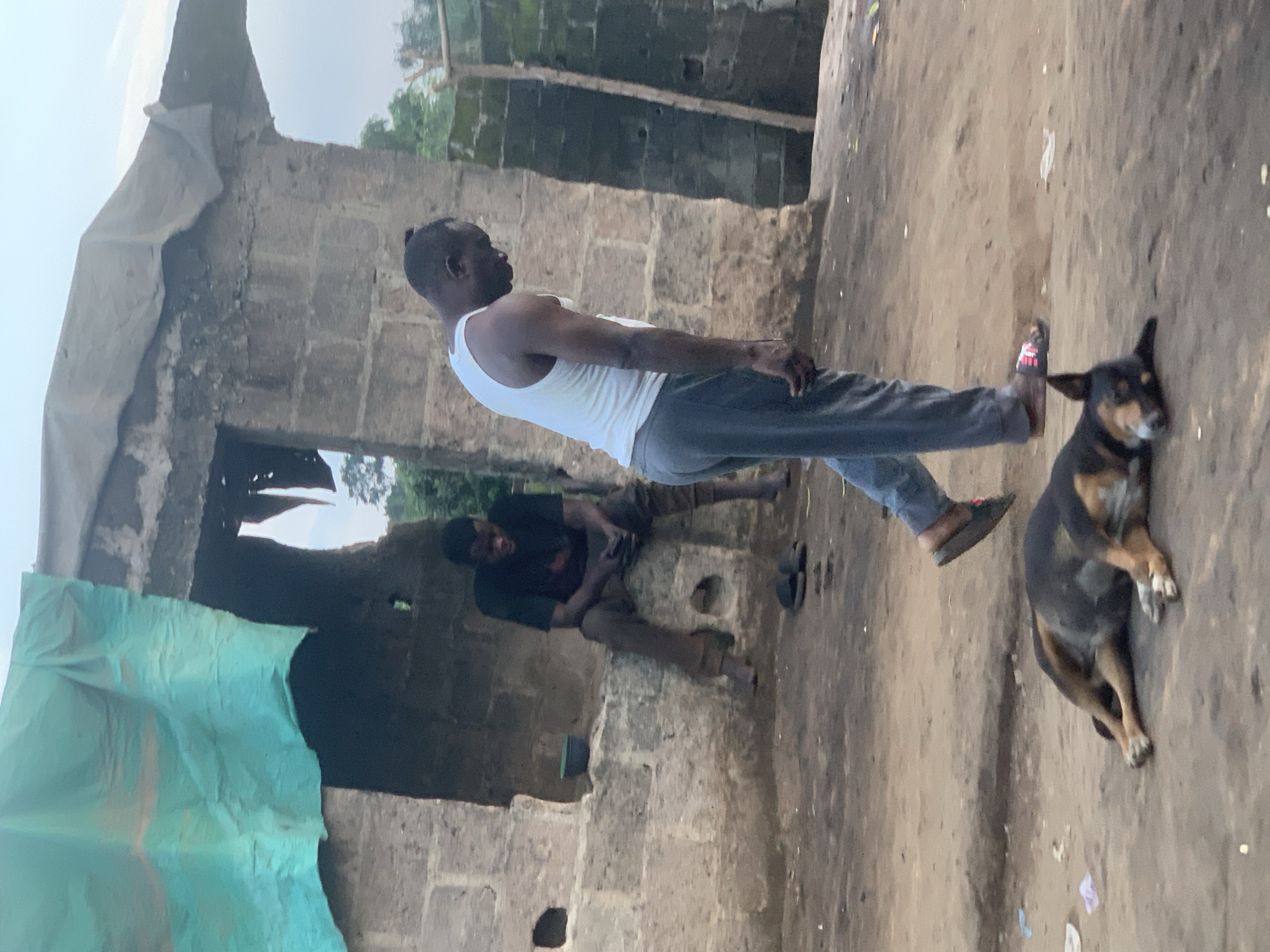 The image depicts an outdoor scene in what appears to be a developing area, likely in Shagamu, Nigeria. In the foreground, a medium-sized dog is lying on the dusty ground, appearing relaxed or asleep. Behind the dog, a man wearing a white tank top and grey sweatpants walks away from the camera, his head bowed. Further in the background, another man is seated within an open, unfinished structure made of concrete blocks. He is wearing a dark t-shirt and brown trousers. The structure has a rough, unfinished roof partially covered with a turquoise tarp. The ground is a mixture of dirt and debris, suggesting a rural or informal urban setting. The overall environment appears dry and unkempt.