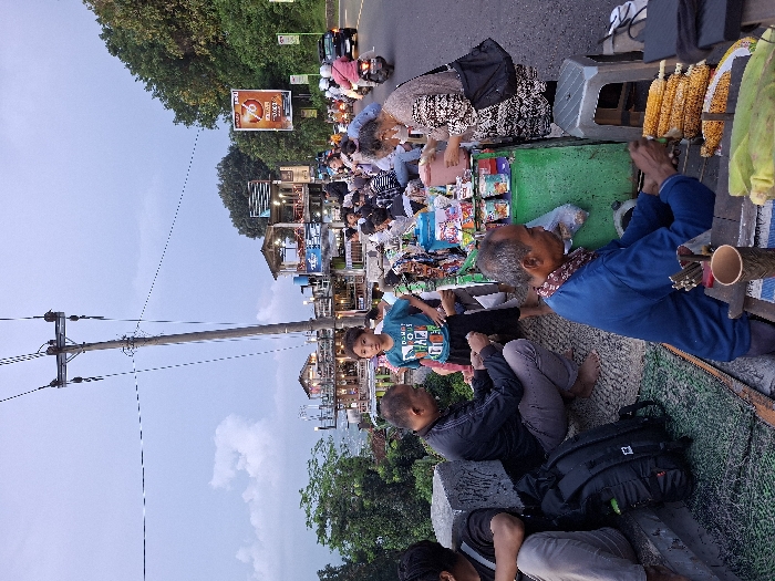 The scene takes place on a street corner in the Kecamatan Piyungan district of Yogyakarta, Indonesia. The coordinates are -7.846 latitude and 110.48 longitude. There are people on the sidewalk in front of a food stand. There are two men seated on the ground in front of the stand, and one man in a blue shirt, seated at a table. In the background there are a few people standing and walking and one man wearing a black shirt who is crouched down. They are all looking at the food stand. The stand has various products on display, and several individuals are seated around it. A young boy in a blue shirt stands to the left of the food stand. The stand is surrounded by a green metal frame. Next to the stand is a gray metal stool. The stand is covered in a black plastic tarp and a green cloth. The road is paved with asphalt. There is a tall power pole with wires running along it. There is a sign on the right of the image. The sign is orange with black lettering. Behind the power pole and the sign is a hillside with green trees growing on it. There is a building visible in the background to the left. The sky is blue with white clouds. The road is empty except for a few parked motorbikes and a car that is driving away. There are trees on the right side of the image, and a sidewalk on the left side. There is a concrete wall in the foreground, and a green mat on the ground.