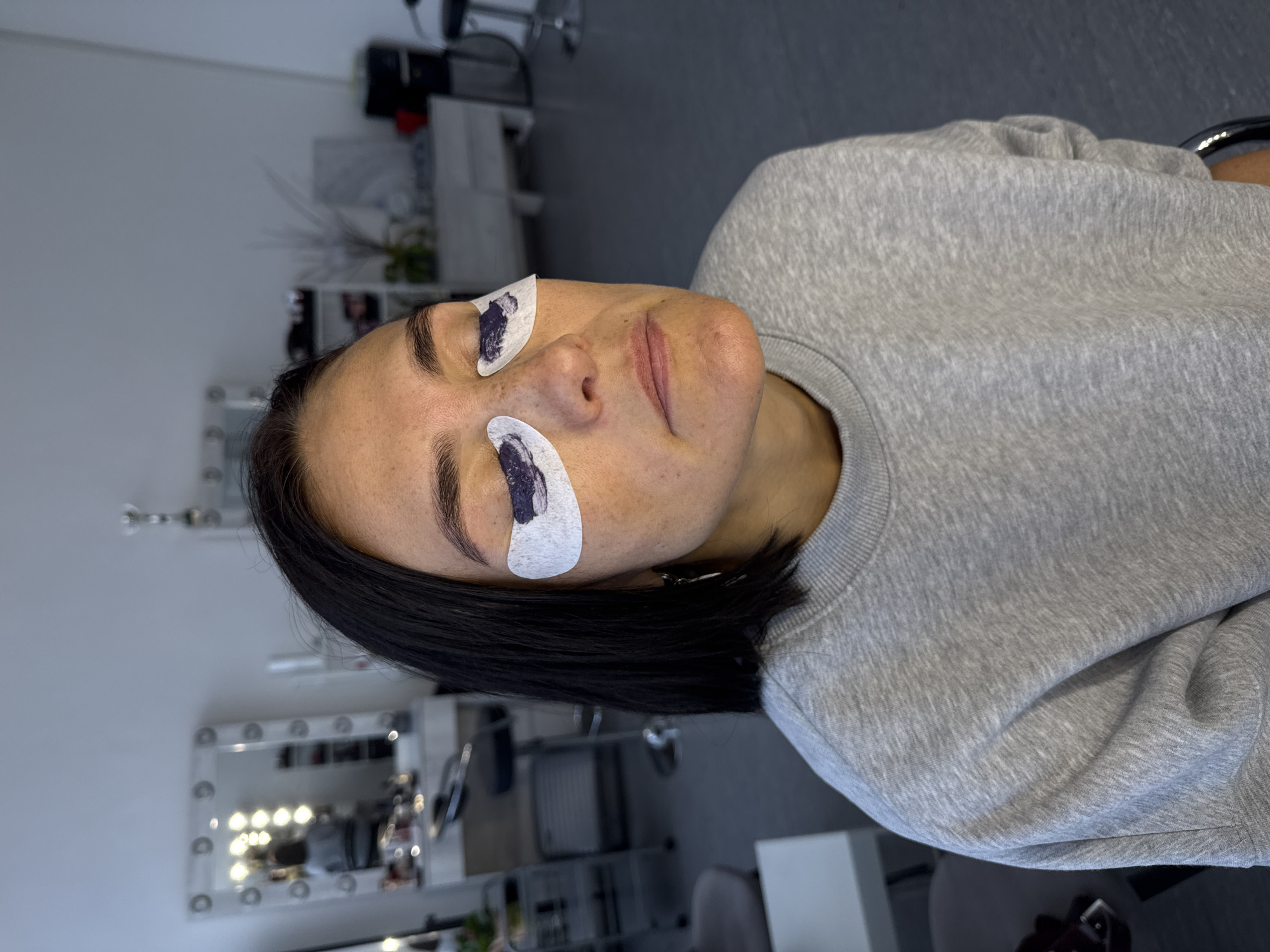 The image depicts a person lying down, with eye patches applied, presumably in a beauty salon setting. The person has dark hair and is wearing a grey sweatshirt. Two white eye patches with dark markings are positioned under each eye. The background reveals elements of a beauty salon, including a lit mirror, chairs, and various beauty supplies. The location is Izhevsk, Russia, based on available information.