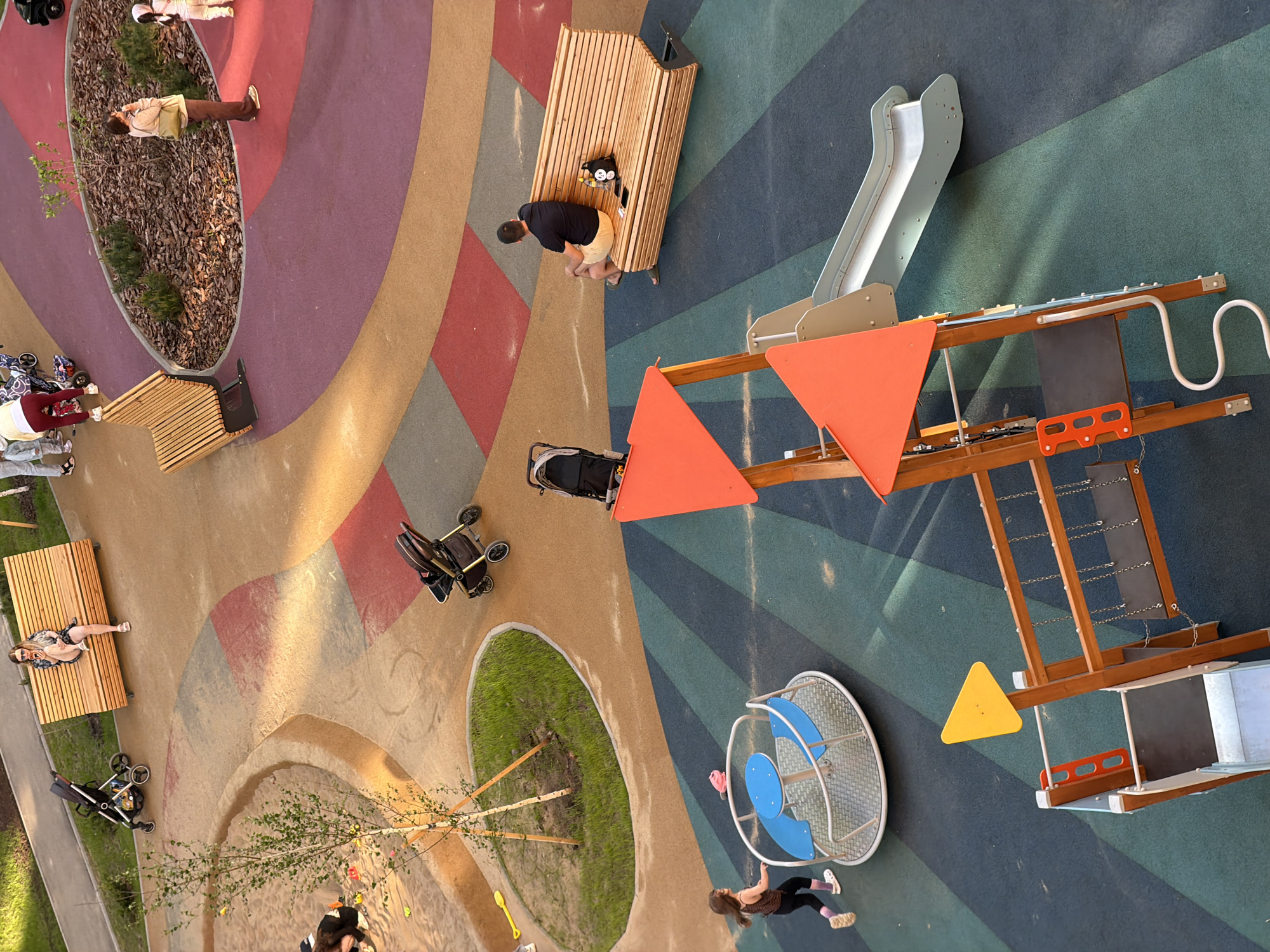 Captured from an aerial perspective, the image shows a playground in Pervomaysky, Russia. The playground features several distinct areas, including a section with colorful patterned ground coverings, a wooden bench area, and a play structure with a slide and red triangular elements. There's also a merry-go-round type of equipment in blue and white. People are present in various locations, including individuals sitting on benches, a person bending over near a wooden structure, and a child interacting with the merry-go-round. Strollers are positioned near the benches. The playground also includes landscaping with grassy areas and a small tree.