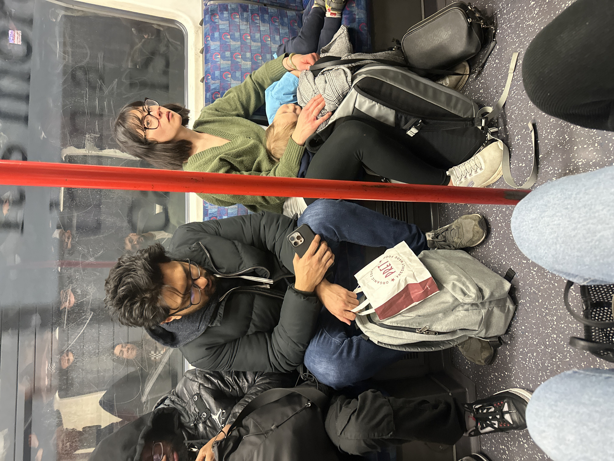 Inside a London Underground train car, a woman with dark hair and glasses, wearing a green sweater, is holding a young child wearing a blue hat. They are sitting near a window, with luggage including a gray backpack and a black purse on the floor nearby. A man with dark hair and glasses, wearing a dark jacket, is seated next to them, holding a phone and a Pret a Manger bag on top of a gray backpack. Other passengers are visible in the background through the window and in the seating area. The train car has a red handrail and blue patterned upholstery. The scene appears crowded.
