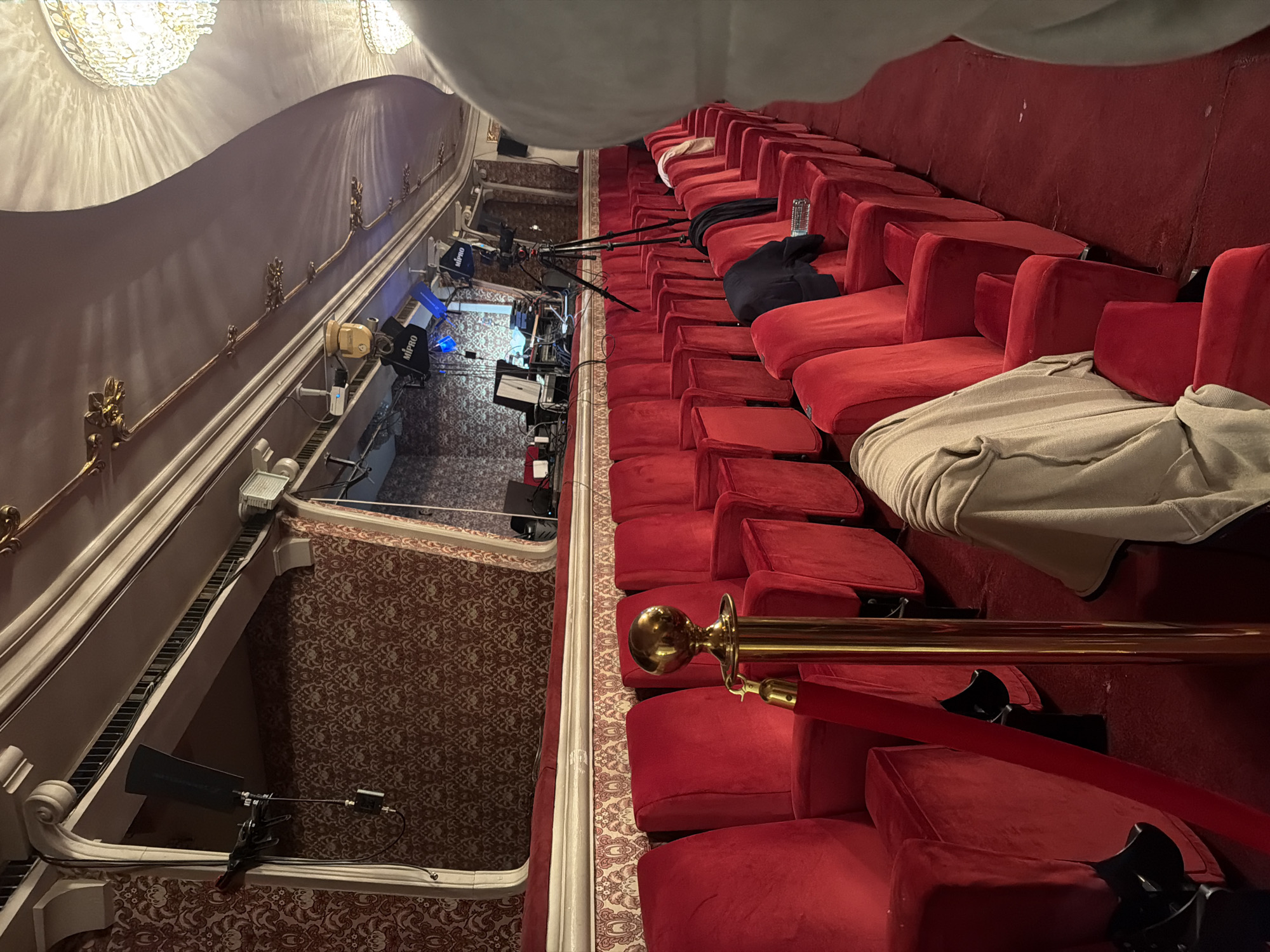 The image displays the interior of an opulent theater or opera house balcony, viewed from an elevated perspective looking towards the stage area. Rows of plush, empty red velvet seats, characteristic of a classic performance venue, dominate the right side of the frame. A shiny gold railing, adorned with decorative finials, runs in front of the seating section, separating it from the lower part of the auditorium.
The wall on the left side of the image features ornate moldings with gold accents, and large, elaborate chandeliers are visible at the top, casting a soft, warm light on the ceiling and walls. The floor is covered with a patterned carpet in shades of red and gold.
Along the edge of the balcony, several pieces of technical equipment are set up on tripods, pointing towards where a stage would be. These include stage lights and audio-visual gear, with the brand name "MIPRO" visible on some of the devices. Cables and other equipment components are scattered around these setups, suggesting ongoing preparations or a recent event.
Personal items are left on some of the red seats: a clear plastic water bottle, a dark jacket, and a light-colored garment (possibly a sweater or blanket). A red velvet rope is also visible, likely used for crowd control or sectioning off seating. The artificial lighting implies an indoor setting, possibly during the evening or night. The scene captures a quiet moment in a grand performance space, with technical arrangements indicating a show is either about to begin, in rehearsal, or has recently concluded.