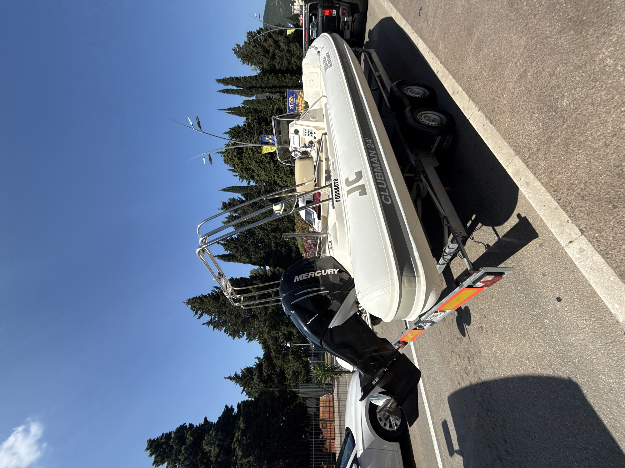 A white rigid inflatable boat, labeled "CLUBMAN 24" and "FOUSKOTO", is parked on a trailer on a paved surface. The boat has a black Mercury outboard motor attached. A white car is parked to the left of the boat, and a black pickup truck is parked to the right. In the background, there are tall evergreen trees and a clear blue sky. The location appears to be a street in Budva, Montenegro, given the presence of Mediterranean cypress trees and the style of the buildings and vehicles.