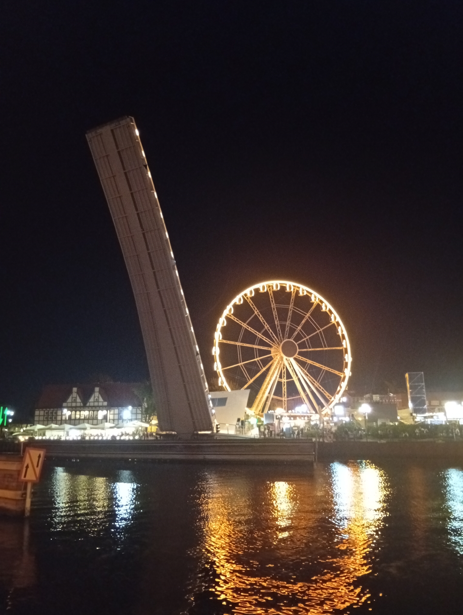 This is a nighttime photograph of the Ołowianka quay in Gdańsk, Poland. The image features a raised drawbridge on the left, its span angled upwards. To the right of the drawbridge, a brightly lit Ferris wheel stands prominently. In the background, illuminated buildings with traditional architecture are visible along the quay, with several white umbrellas indicating outdoor seating. The dark water in the foreground reflects the lights from the Ferris wheel and the buildings, creating a shimmering effect. A sign with two upward-pointing arrows is visible near the water's edge on the left.