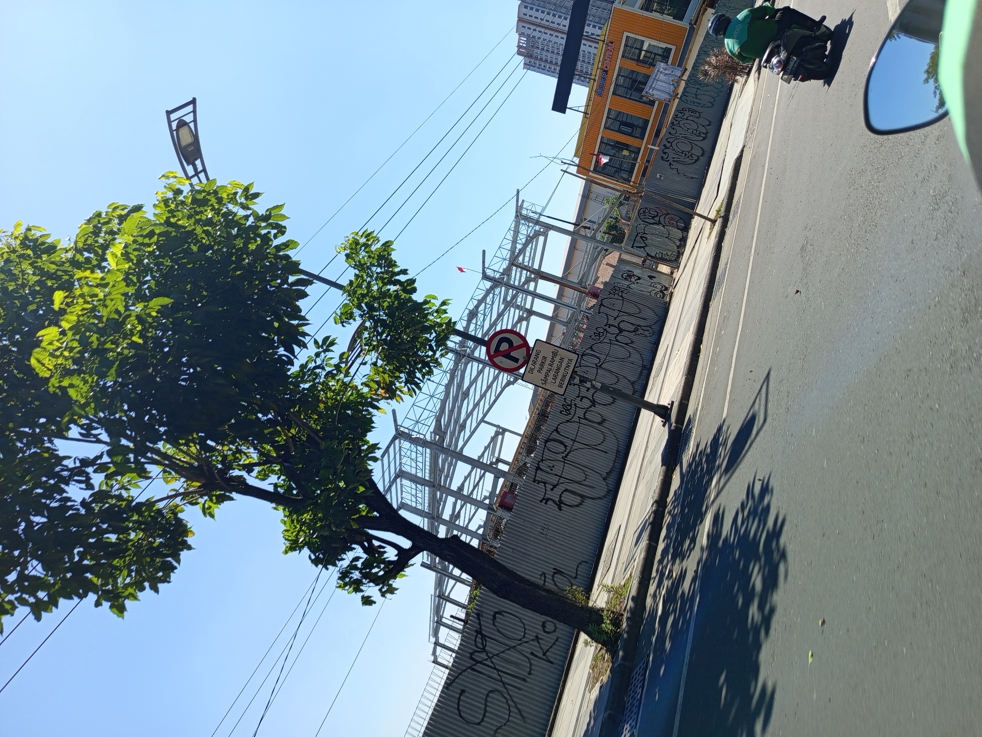 The image depicts a street scene in Surabaya, Indonesia, on a clear, sunny day. In the foreground, a motorcycle is seen from behind, with its rider wearing a green jacket. The motorcycle is traveling on a paved road. To the left, a young tree with lush green leaves casts a shadow on the road. In the background, a construction site is visible, enclosed by a corrugated metal fence covered in graffiti. A metal frame structure for a building is being erected on the site. A street lamp with a modern design is mounted on a pole to the right of the tree, partially obscured by its branches. Further in the background, an orange building with "WARCOP CENTRAL" written on it is present. To the far right, a tall, modern building stands against the blue sky. A sign with a "P" in a circle with a diagonal line through it, indicating "no parking," is posted on a pole in front of the construction site. The overall atmosphere is one of urban activity with ongoing development and daily life.