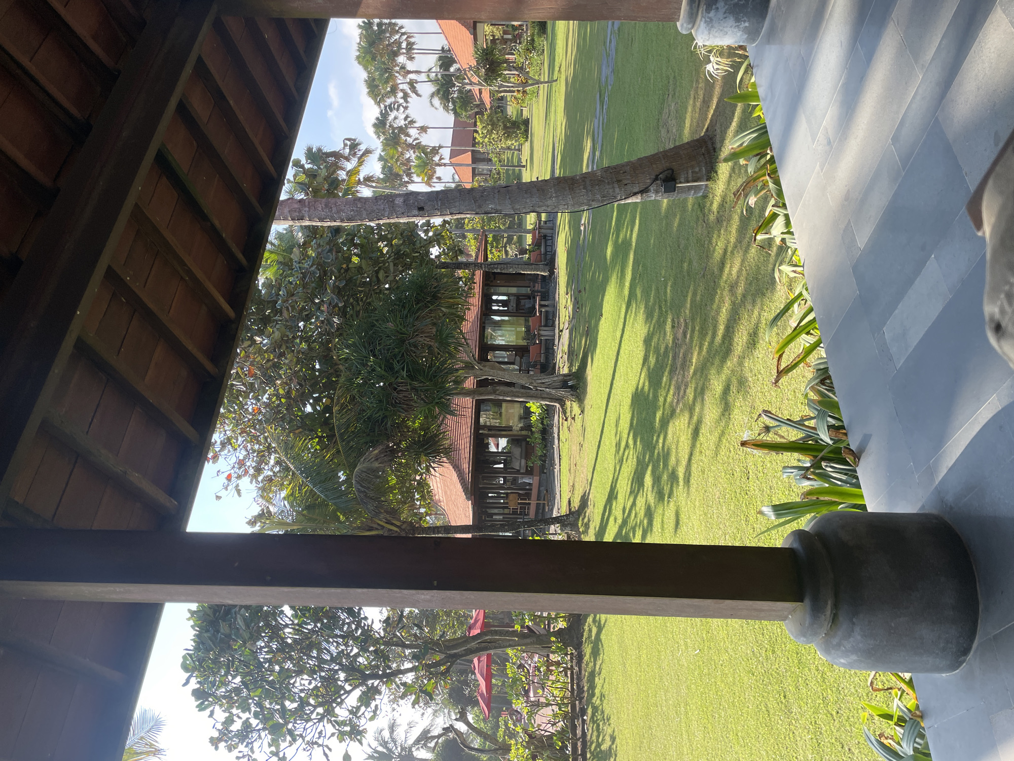 This image depicts an outdoor scene at a resort in Selemadeg Timur, Indonesia, likely during the daytime. The viewpoint is from under a wooden-roofed structure, looking out onto a lush green lawn. In the foreground, a thick, dark wooden pillar with a rounded stone base is visible on the left, partially obscuring the view.
The middle ground features a grassy expanse with scattered trees, including palm trees. In the center of the frame, a low-rise building with a red tile roof and open-air seating is visible. Several other similar buildings with red roofs are scattered across the property in the background. Red umbrellas are also visible on the lawn to the left. The environment appears well-maintained and tropical, with ample sunlight casting shadows on the grass. No people are discernible in the image.