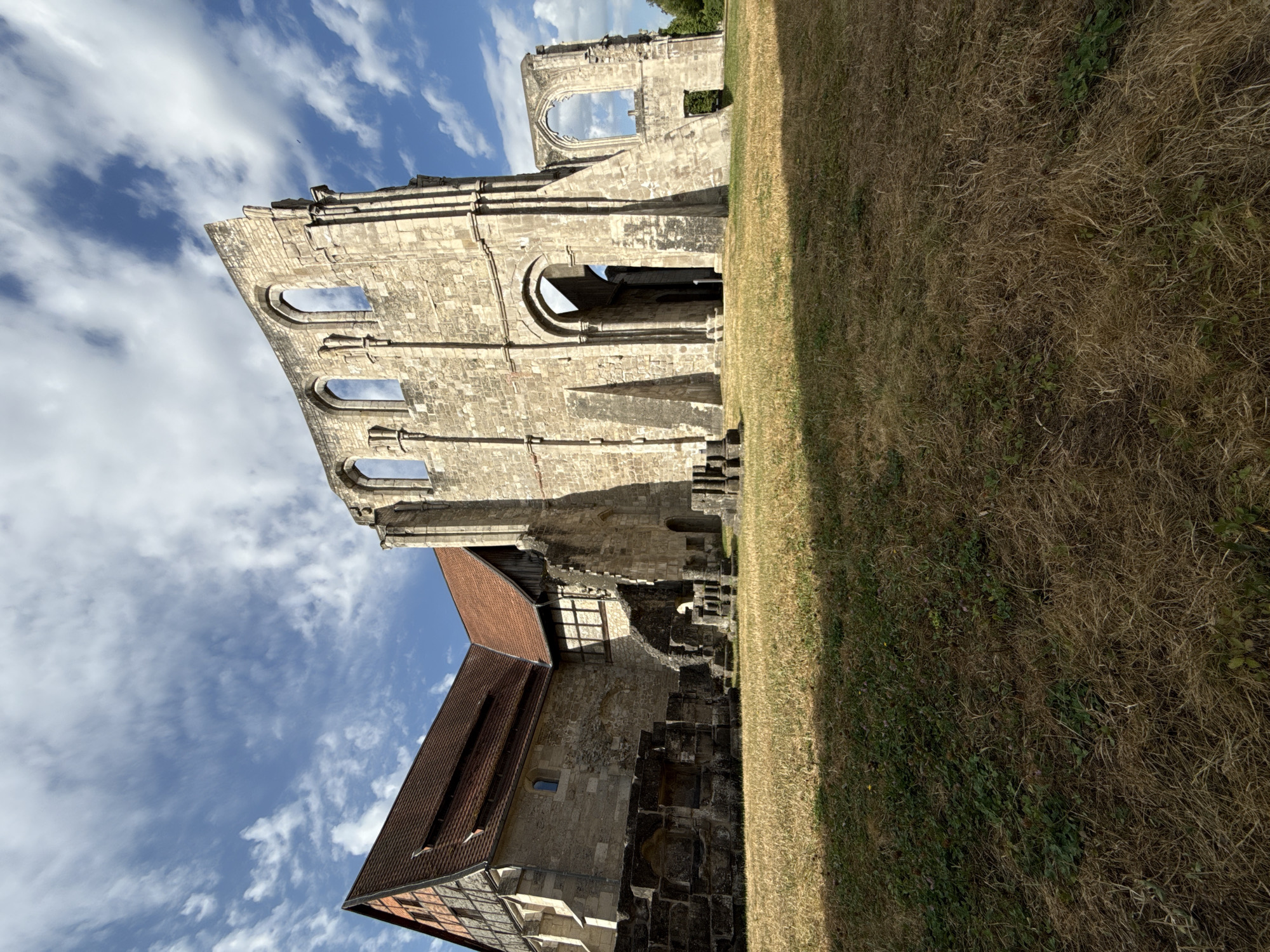 The image depicts the ruins of the Walkenried Abbey in Walkenried, Germany, under a partly cloudy sky. The foreground is dominated by dry, brown grass. To the left, a portion of the preserved abbey building stands with a red-tiled roof and half-timbered elements. Adjacent to this, the stone ruins of the abbey rise, characterized by arched windows and weathered stone walls. The ruins show significant decay, with missing sections of walls and roofs. The background features a clear blue sky with scattered white clouds. The overall scene conveys a sense of historical grandeur and the passage of time.