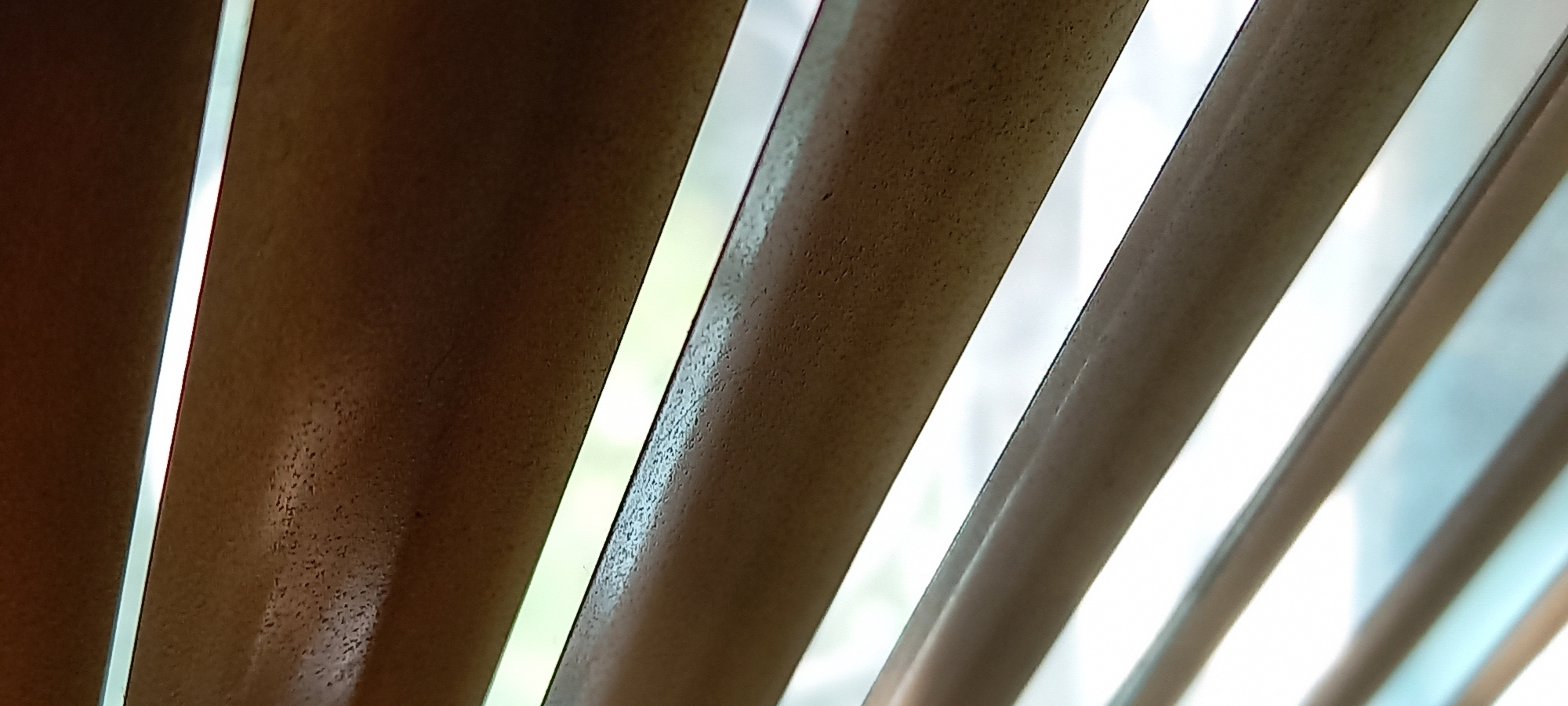 The image provides a close-up, abstract view of horizontal window blinds, likely made of wood or a wood-effect material, angled diagonally across the frame. The slats are a dark, warm brown color and exhibit a textured surface, appearing slightly coarse with numerous small, dark speckles.
Bright, narrow strips of light are visible through the gaps between the partially opened slats, indicating an exterior environment beyond. The light filtering through is predominantly bright white, though some areas show a faint greenish or bluish hue, suggesting the presence of outdoor foliage or the sky. The intensity of the light confirms it is daytime.
The perspective is from below, looking upwards and outwards through the blinds, emphasizing the strong diagonal lines formed by the slats and the shafts of light. No people, specific objects, or activities are discernible within the frame. There is no visible text. The scene is static, capturing the interplay of light and shadow created by the blinds in an interior setting in Kryvyi Rih, Ukraine.