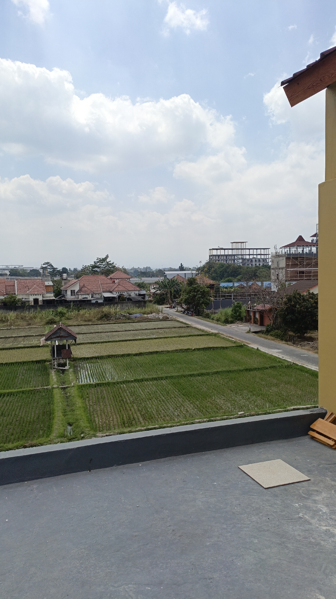 The image depicts a view of rice paddies in Trihanggo, Indonesia, from an elevated perspective. In the foreground, a gray concrete surface with a single square tile is visible, bordered by a dark gray railing. Beyond the railing, lush green rice fields are arranged in terraces, with a small wooden hut situated in the middle of the paddies. A dirt road runs alongside the fields, with several motorcycles parked near it. In the background, residential houses with red tile roofs are nestled among trees. Further in the distance, an unfinished multi-story building under construction is visible, along with other structures that appear to be part of a developing area. The sky is partly cloudy with a light blue hue.