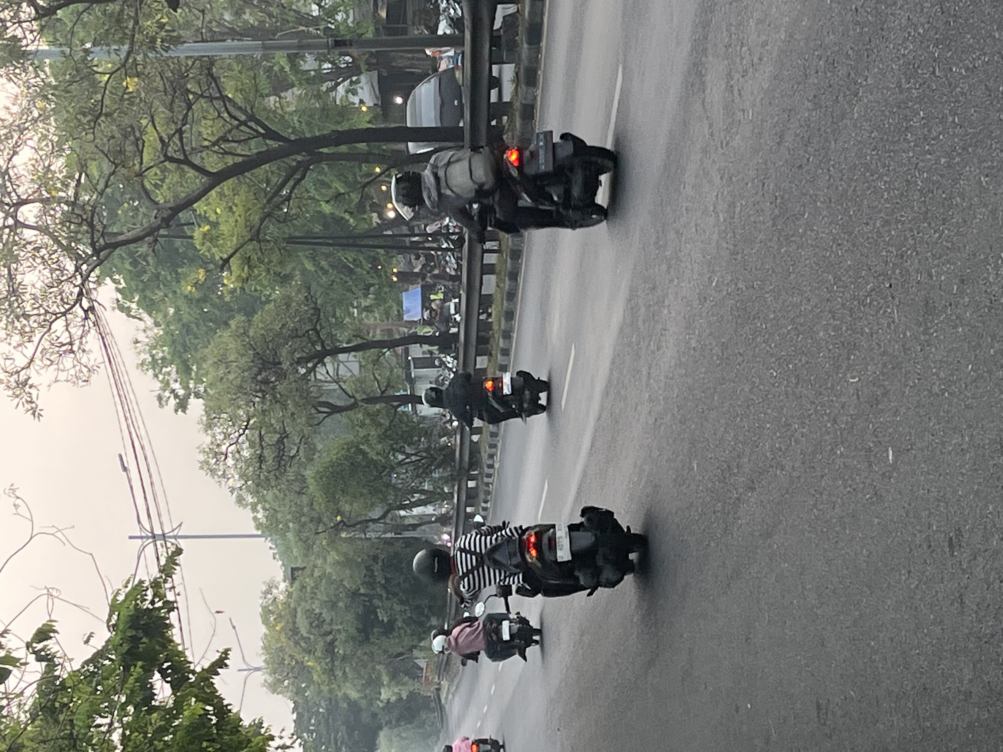The image captures a street scene in Kludan, Indonesia, featuring several individuals riding motorcycles. In the foreground, a person wearing a black and white striped shirt and a helmet is riding a dark-colored scooter. To the left, another motorcyclist in a pink jacket and white helmet is visible. Further ahead, two more motorcyclists are seen, one in black attire and a helmet, and another also in dark clothing with a backpack. The motorcycles are traveling on a paved road bordered by trees and a metal railing. In the background, there are more trees, power lines, and parked vehicles. The sky appears overcast, suggesting either early morning or late afternoon.