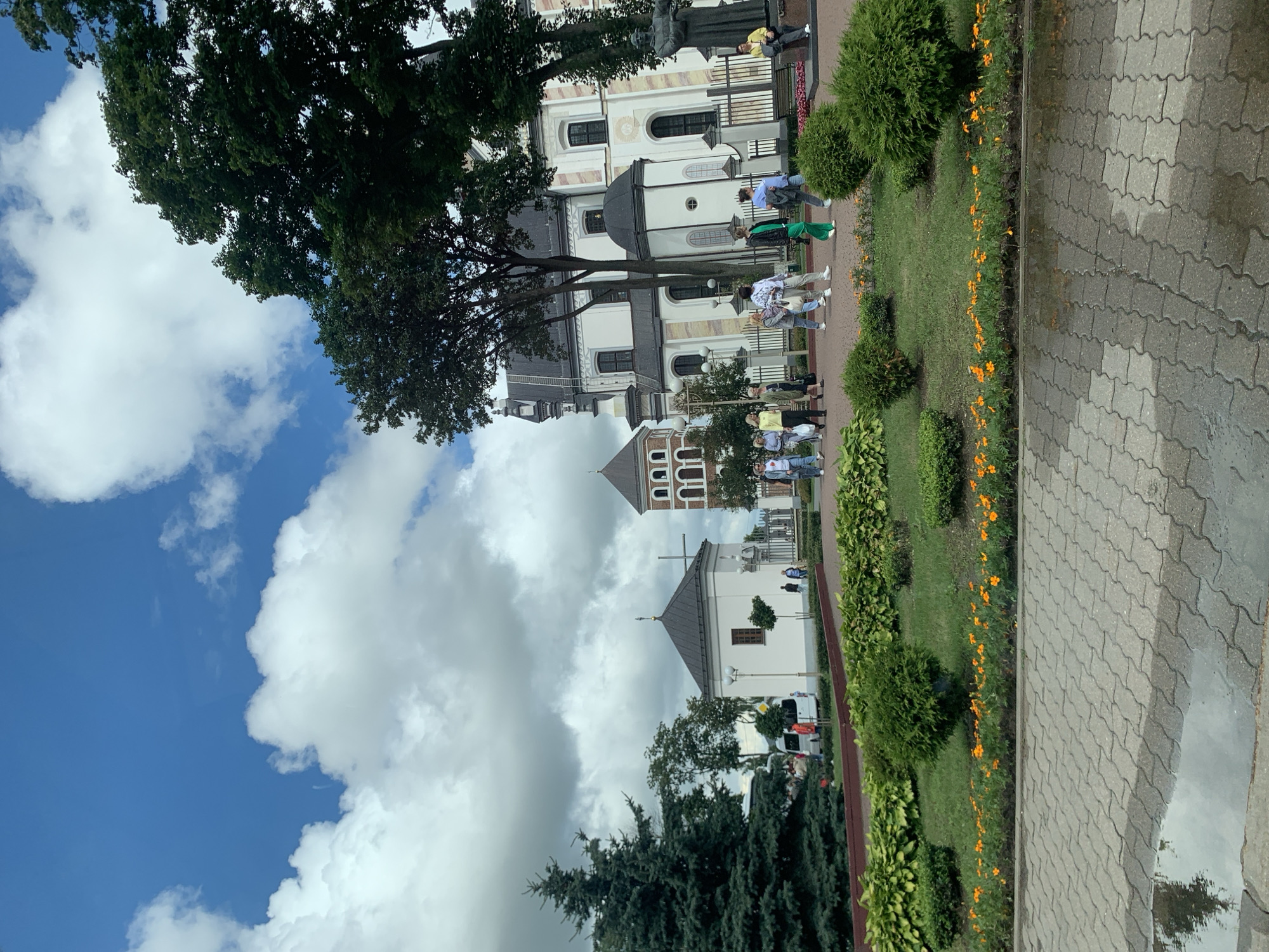 The image depicts the Niasvizh Castle complex in Niasvizh, Belarus, under a partly cloudy sky. In the foreground, a paved walkway with a puddle reflects the scene. A manicured garden with trimmed bushes and a line of small orange flowers separates the walkway from a grassy area. Several people are walking along a path in front of the castle complex. The castle itself is a large, white building with a dark roof, featuring numerous windows and architectural details. Adjacent to the main building is a smaller, white structure with a dark roof and a cross, and further to the left, a taller, brick tower with arched windows. Green trees, including a prominent evergreen on the left, are scattered throughout the scene. A statue of a man in a seated position is visible on the right side of the image. Several vehicles, including a white van, are parked in the background. The overall scene is that of a historical and cultural landmark with visitors enjoying the grounds.