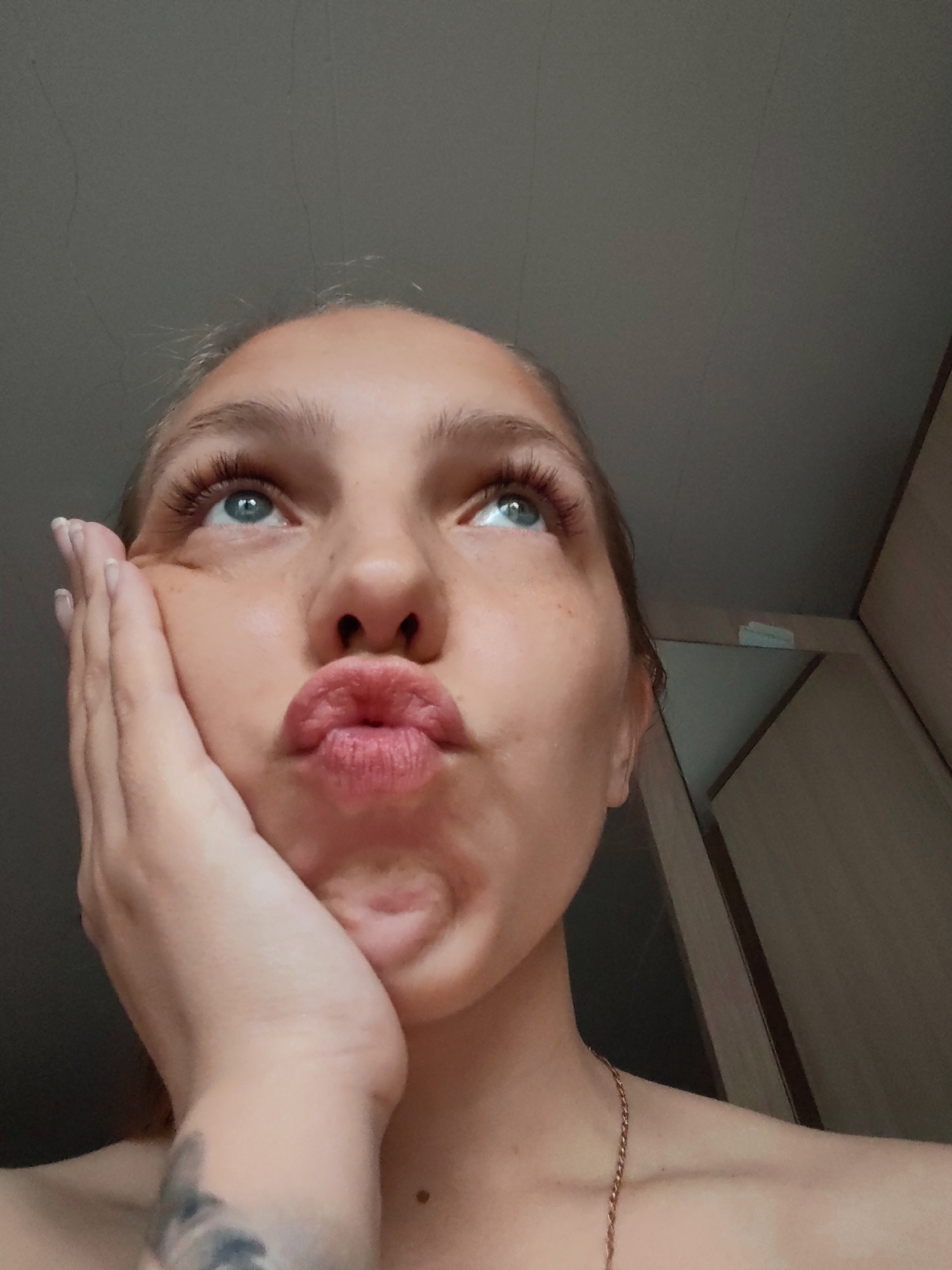 A close-up, low-angle portrait features a young female looking upwards and to the right of the frame. Her head is slightly tilted back, revealing her light skin tone and what appears to be light blonde or light brown hair around her forehead. Her eyes, a light blue or green, are open wide, gazing intently upwards. Her eyelashes are prominent, and faint freckles are visible on her nose and cheeks. Her lips are pursed into a "duck face" or kissy expression.
Her right hand is pressing against her right cheek and chin, with her fingers slightly spread. Her fingernails are neatly manicured with a light-colored polish. A portion of her left shoulder and collarbone is visible, adorned with a thin gold chain necklace. On her left forearm, a section of a tattoo featuring dark, abstract, or possibly floral designs can be seen.
The background consists of a plain, light grey or beige ceiling or wall, indicating an indoor setting. On the right side, part of a light brown wooden cabinet or door is visible. The lighting is diffused and soft, without strong shadows, suggesting indoor illumination. The overall impression is a casual, posed photograph, possibly a selfie, conveying a playful or contemplative mood. The scene is set indoors, within a residential environment in Chaikovsky, Russia. There is no visible text in the image.