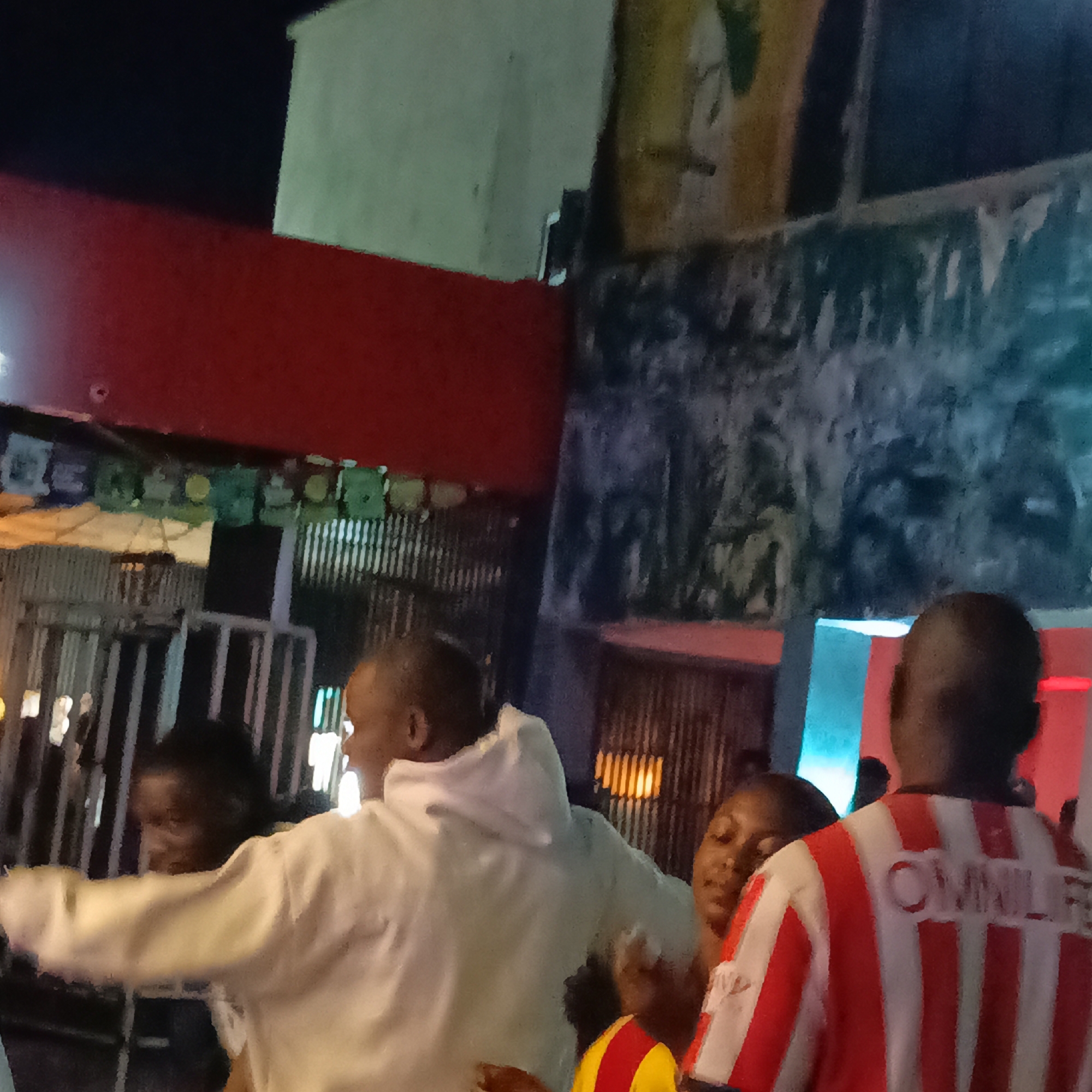A dimly lit outdoor scene in Akwanga, Nigeria, possibly at night. Several people are visible, including a person in a white hoodie with outstretched arms, a person wearing a red and white striped shirt with "OMNILE" printed on the back, and another person wearing a yellow and red shirt. The background features a mix of structures including corrugated metal and painted surfaces with graffiti or other artwork. Decorations in green, white, and blue appear to be hanging overhead near the front. A metal gate or barrier is visible in the middle ground, and there are signs of lighting or illumination from multiple sources. The overall setting seems to be a gathering or event outdoors.