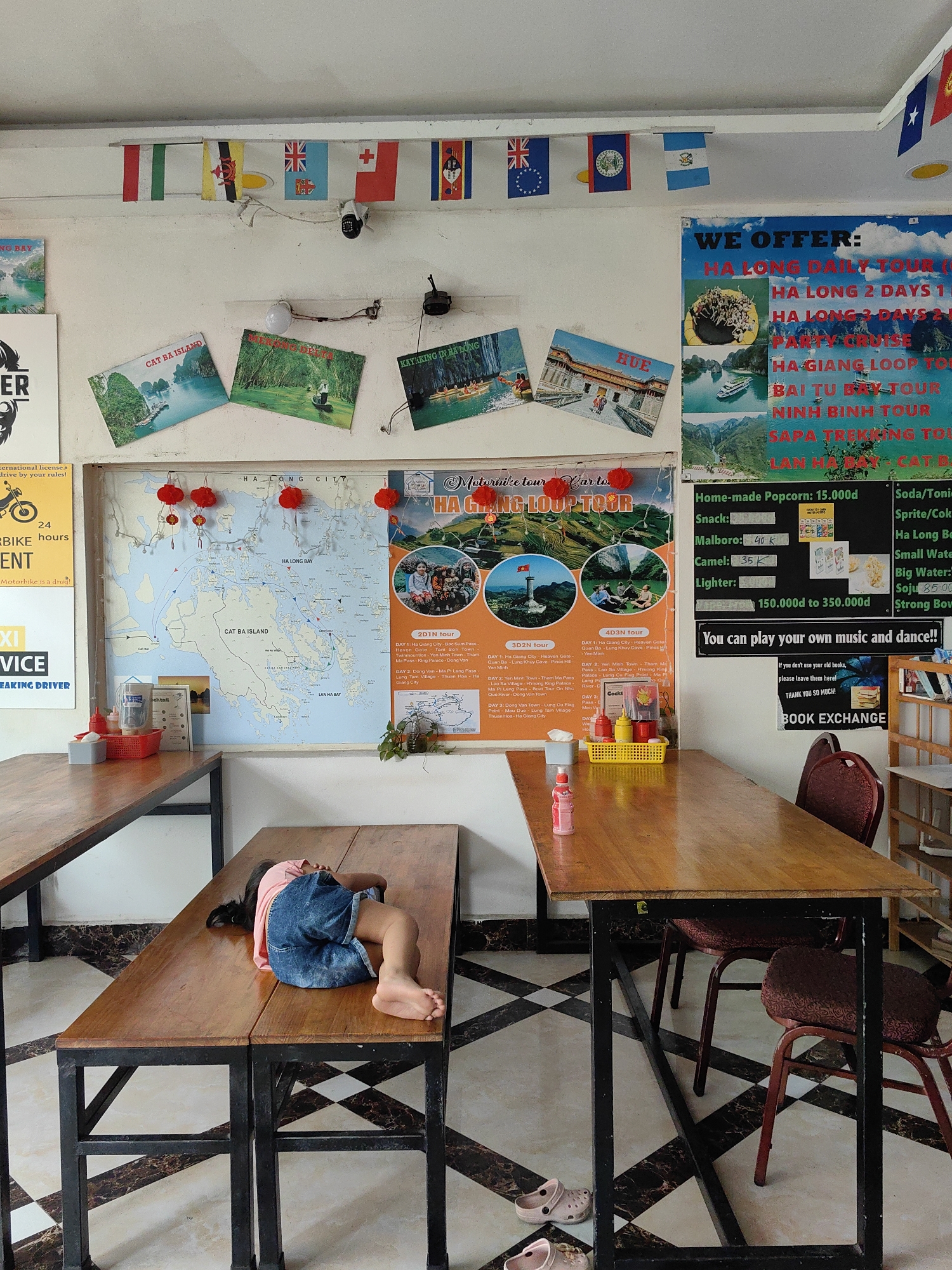 A young child sleeps on a wooden bench in a brightly lit room in Bai Chay Ward, Vietnam. The room appears to be a travel agency or tourist information center. Various flags hang near the ceiling, and several posters and maps adorn the walls, advertising tours of Ha Long Bay and other local attractions. A table with two chairs is situated near the bench where the child is sleeping. A pair of children's shoes lies on the floor next to the table. The floor has a black and white checkered pattern.