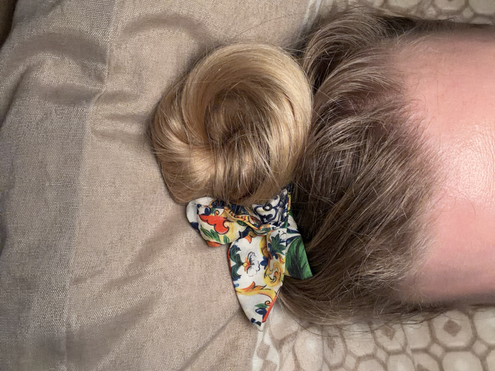 A close-up, top-down view depicts the upper portion of a person's head. The subject's hair, light brown at the roots, transitions to a lighter blonde shade, gathered into a coiled bun atop the head. This bun is secured by a scrunchie featuring a white background with a vibrant, multi-colored floral or abstract design in red, orange, yellow, blue, and green. The visible forehead and upper temples display a light skin tone with reddish undertones. The subject lies on a soft, textured, light beige fabric surface, possibly bedding, exhibiting folds and a fine weave. A portion of a second beige fabric, featuring a repeating geometric pattern, is visible to the lower left. No specific actions or interactions are depicted. The presence of the subject in the city of Catania, Italy, cannot be confirmed from the visual information provided within the image.