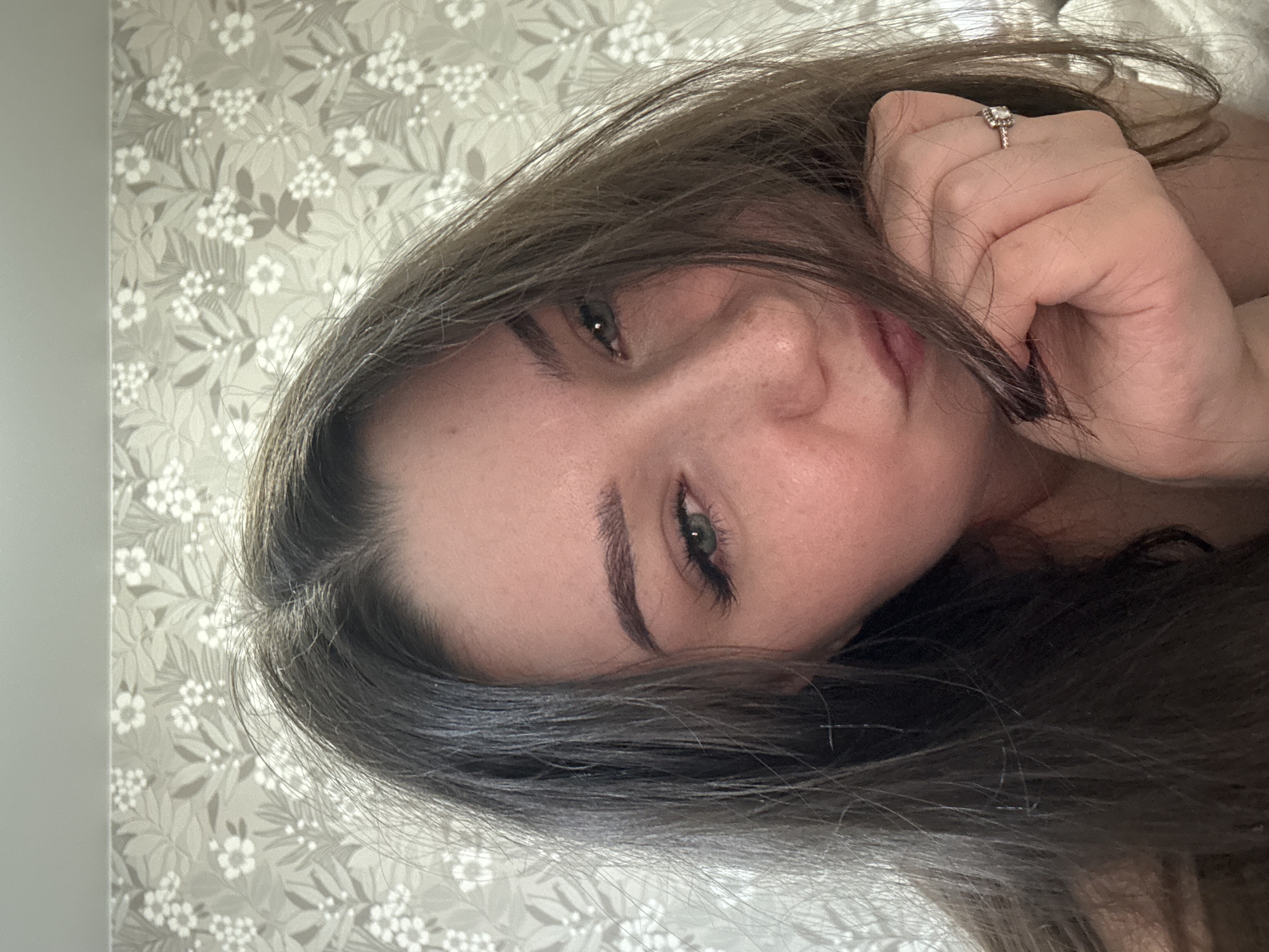 This media file is a close-up, sideways portrait of a young woman lying down, with her head tilted. She has long, dark brown hair that is partially obscuring her face. Her eyes are visible, with dark eyebrows and mascara. She is wearing a ring on her right hand, which is held near her face. The background is a light-colored wallpaper with a floral pattern of white flowers and green leaves. The lighting suggests it might be daytime, possibly indoors, given the presence of wallpaper. The woman's pose and expression are relaxed, suggesting a casual or private moment. There is no visible text in the image. The location context is Zarechnaya (Gamovskoe S/P), Russia.