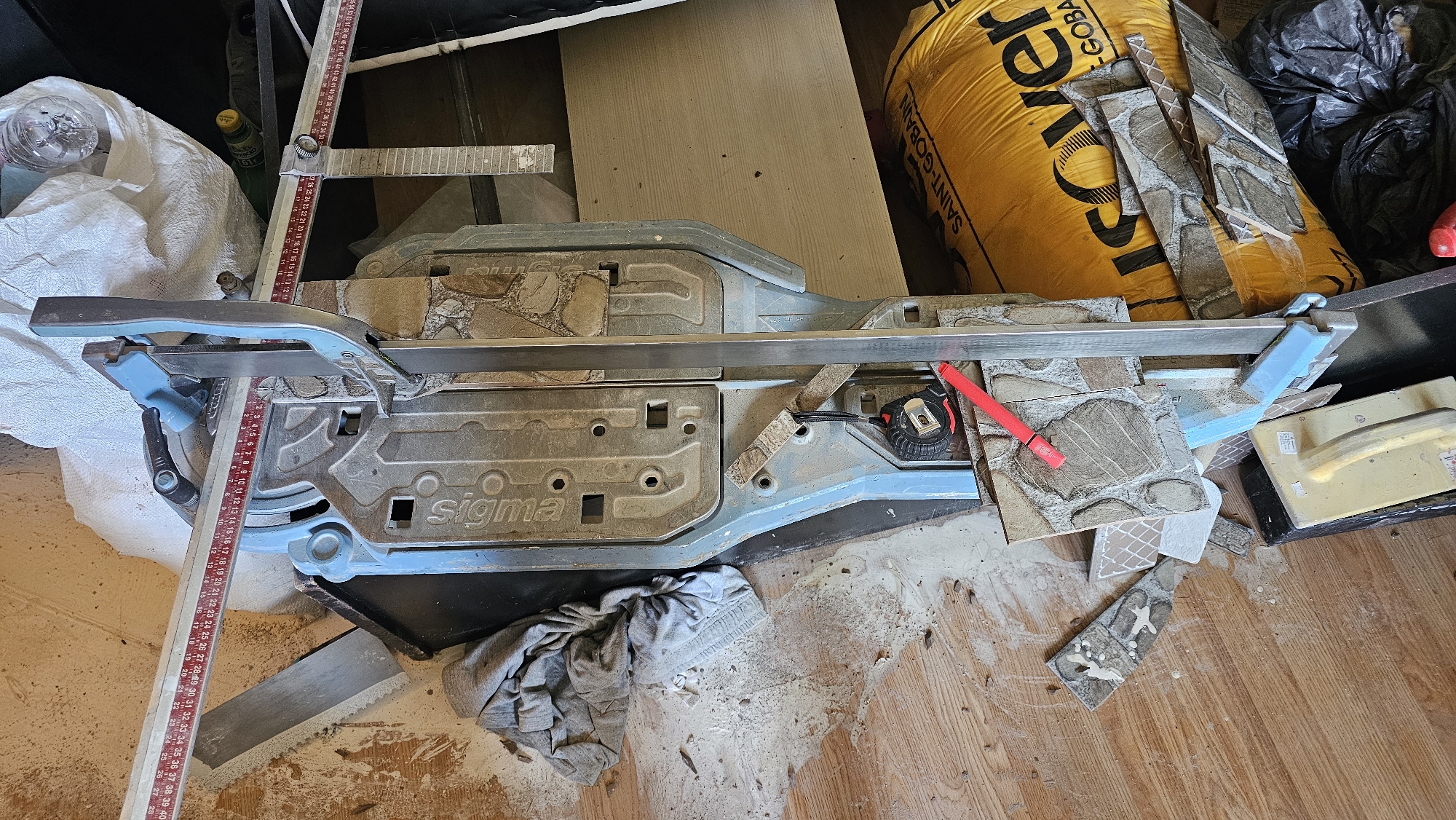 The image shows a tile cutter, likely in a state of disarray, on a wooden floor. The tile cutter is blue and silver, with a long metal bar and a measuring tape attached. Several pieces of tile, some broken, are scattered around the cutter. There is also a yellow bag with black lettering, a grey cloth, and a black garbage bag visible in the background. The overall scene suggests a construction or renovation project that is either in progress or has recently been completed. The location of Sofia, Bulgaria is not indicated by any visual cues in the image.