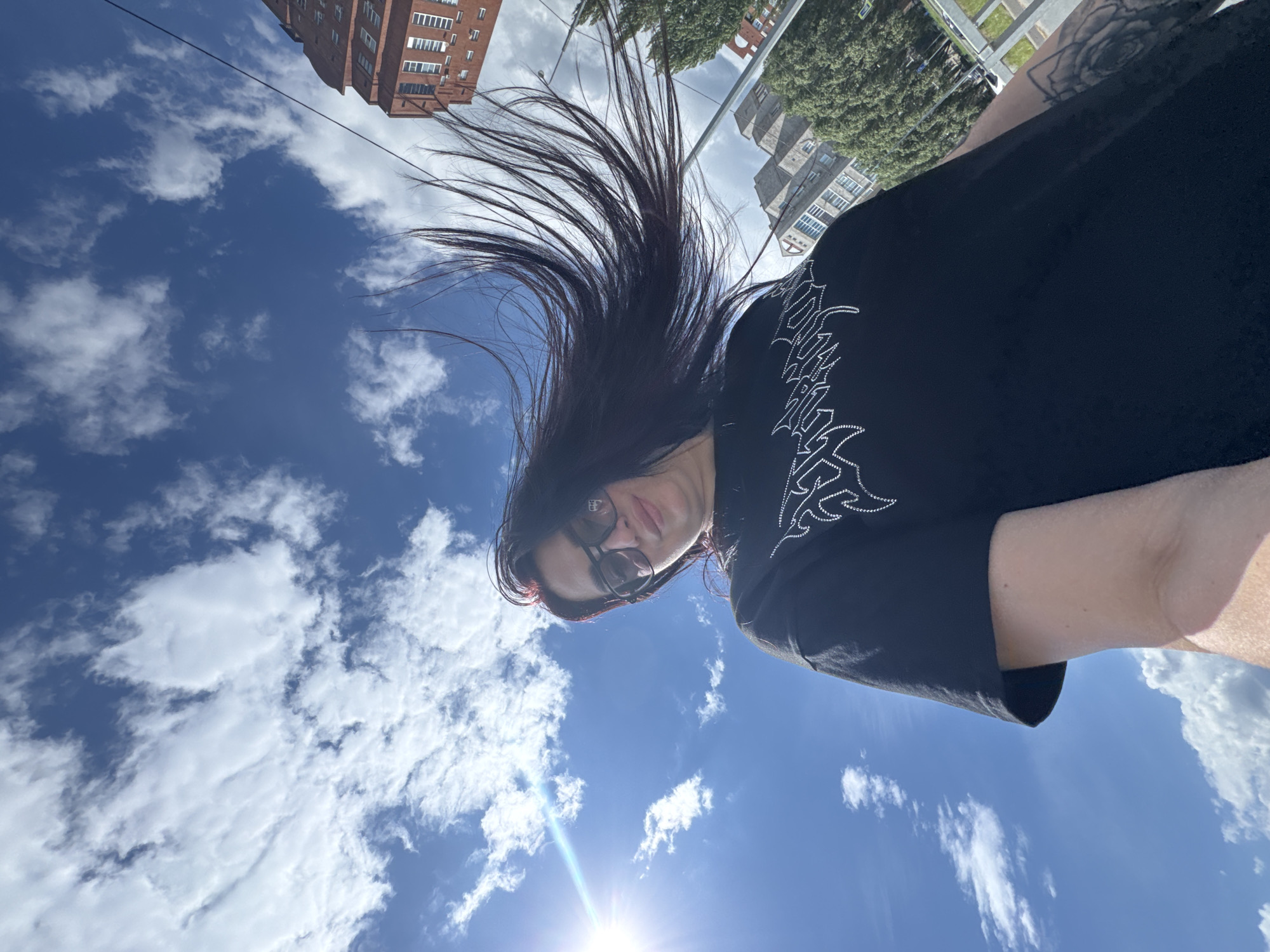 A low-angle, full-body outdoor shot features a woman with long, dark hair styled with dark streaks, wearing dark-rimmed glasses and a black t-shirt with a white graphic. Her hair is blowing to the right, creating a dynamic effect against a bright blue sky filled with fluffy white clouds. The sun is visible on the left side of the frame, casting a lens flare. In the background, several buildings of varying architectural styles are visible, including a tall, reddish-brown brick building and other gray buildings, interspersed with trees and streetlights, suggesting an urban environment. The overall scene is a selfie taken outdoors on a sunny day.