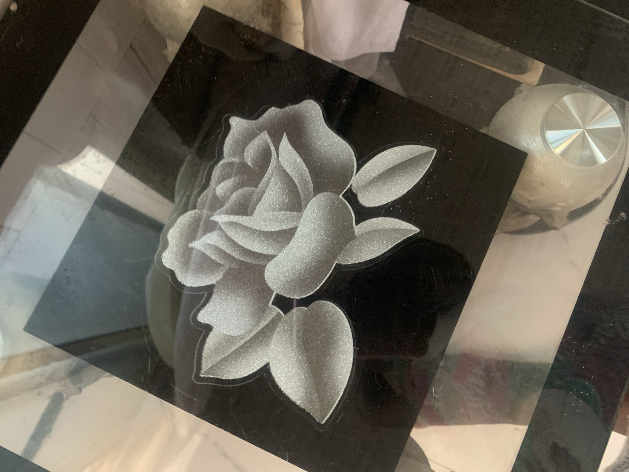 The image shows a close-up, angled view of a black, rectangular surface with a frosted or etched design of a rose with leaves. The rose is rendered in shades of white and light grey, with a shimmering or glitter-like texture. The background is dark and reflective, showing distortions of the surrounding environment. The surface appears to be part of a larger object, possibly a table or a plaque, with parts of reflective, possibly glass or polished metal surfaces visible at the top and right edges. The bottom right portion of the image shows a metallic, circular object with radial lines, possibly a knob or handle, and a glimpse of what might be wooden or metal legs of furniture. The overall impression is one of decorative engraving on a dark, smooth material. The image does not contain enough information to determine its location in the city of Jalingo, Nigeria, or any specific details about people, actions, or interactions.