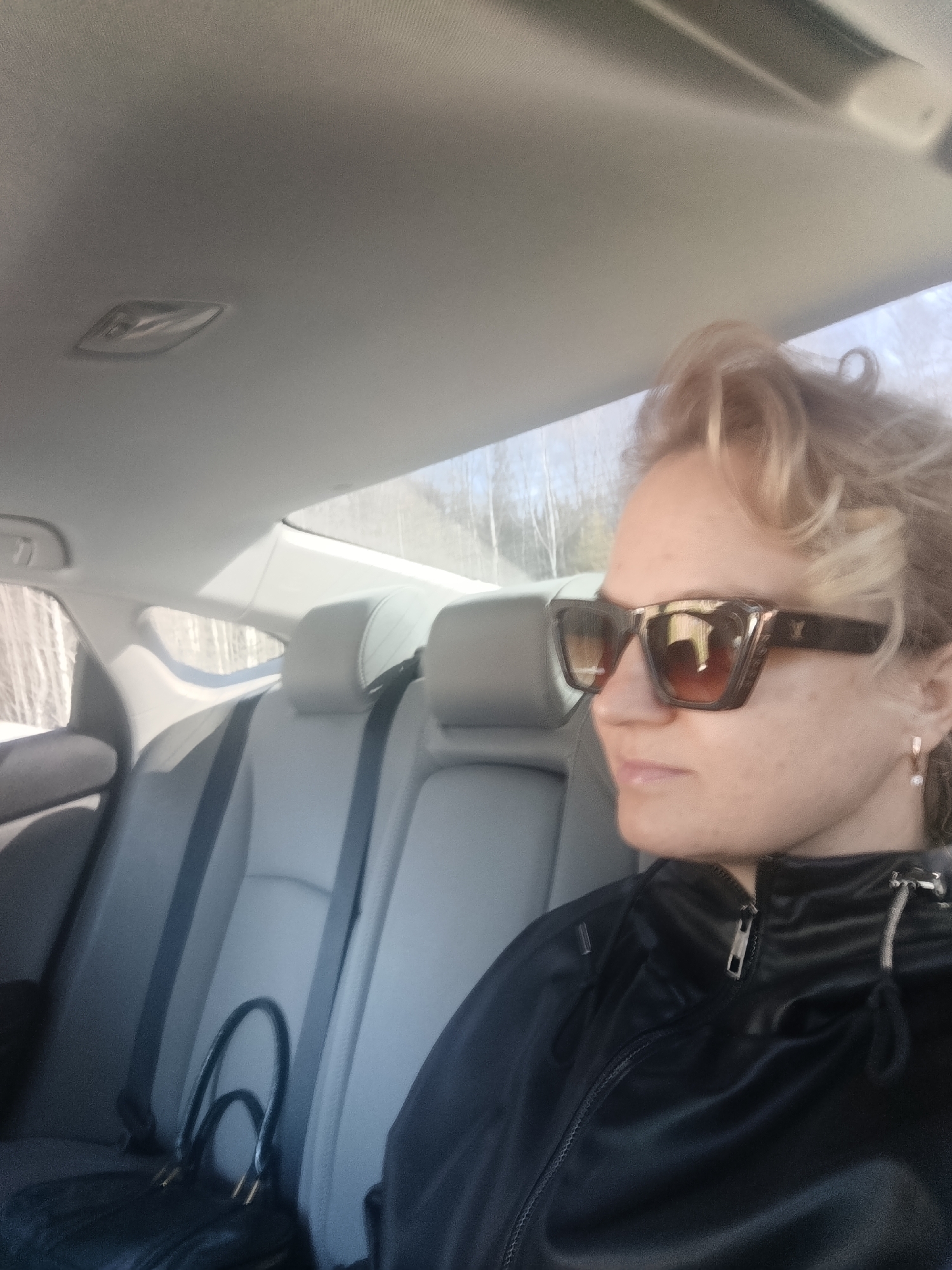 The image depicts the interior of a car in the city of Марковский, Russia, featuring a female individual seated in profile, looking towards the right. She has light-colored hair, wears large, square-framed sunglasses with reflective dark lenses and a decorative upper frame accent, and a black zippered jacket. A small, clear-stone earring is visible on her right ear. A black handbag rests on the left rear passenger seat. The car's interior consists of light grey upholstered seats and headrests. Through the vehicle's windows, a blurred outdoor environment is visible, showing numerous bare trees, suggesting a natural, possibly forested, setting under a bright sky.