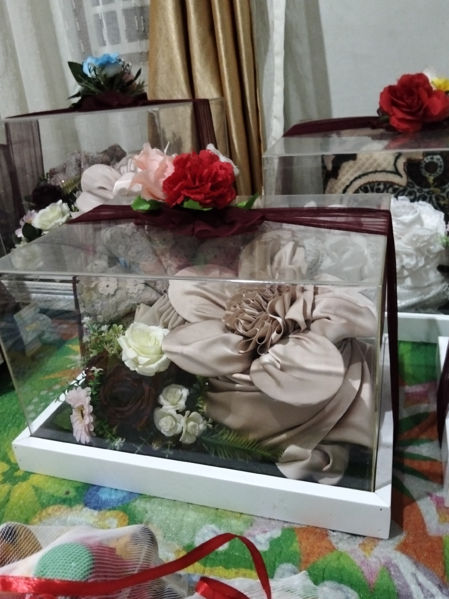 The image displays a close-up of several rectangular, transparent boxes containing fabric gifts, likely for a traditional Indonesian ceremony such as a wedding or engagement. These boxes, decorated with burgundy ribbon and artificial flowers, are arranged on a patterned rug.
In the foreground, a box is filled with folded beige fabric, accented by a floral arrangement of white and dark red roses, and smaller white flowers with green foliage. The box itself rests on a white base. Behind it, another similar box is partially visible, also containing fabric and a floral embellishment. Further back, a third box is visible, showing patterned fabric inside.
To the left, a box with a blue flower arrangement sits atop another box. To the right, a box with a prominent red rose and yellow flowers is visible. The background features gold curtains and a window. The overall impression is one of preparation for a significant event, with the gifts meticulously presented. The lighting suggests an indoor setting, possibly during daylight hours given the brightness. The scene evokes a sense of anticipation and celebration. There is no visible text in the image.
