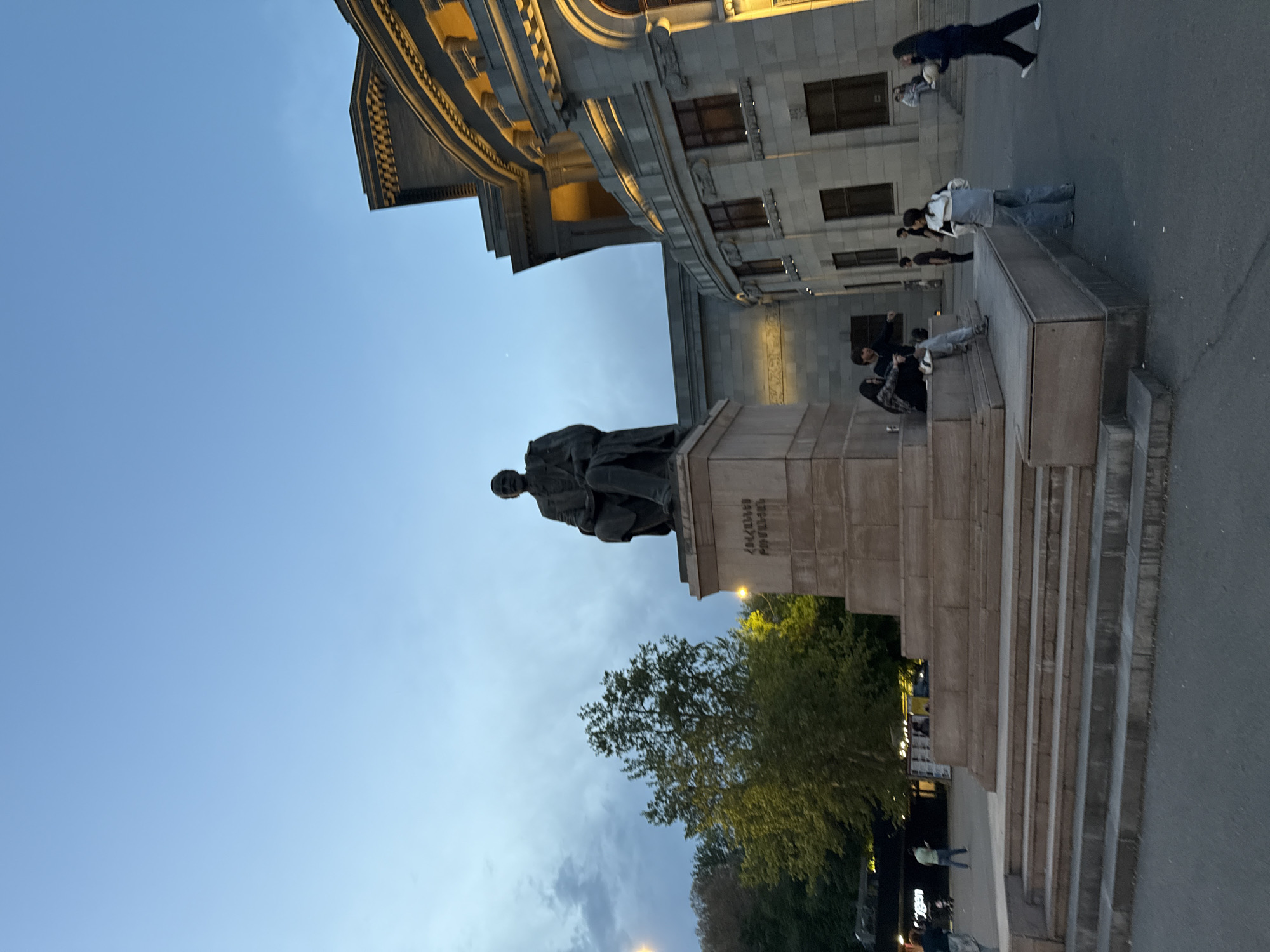 This image depicts a public square in Yerevan, Armenia, captured during dusk or early evening. The scene is dominated by a grand architectural building in the background and a prominent statue in the mid-ground.
The central subject is a bronze statue of a seated male figure, placed on a tall, multi-tiered stone pedestal. Inscriptions in Armenian script are visible on the pedestal, clearly identifying the figure as "Ա. ԻՍԱՀԱԿՅԱՆ" (A. Isahakyan) with the dates "1875-1957," indicating it is a monument to the Armenian poet Avetik Isahakyan.
Behind the statue, a large, ornate building with classical architectural styling forms the backdrop. It features multiple stories, elaborate cornices, arched windows, and decorative stone work, some sections appearing to be lit by warm interior or exterior lights, contrasting with the fading natural light.
Several people are scattered throughout the public space. On the right side, a person in dark clothing is seen walking. Closer to the statue and to the right, a small group of people are relaxing on stone steps or benches, with some seated and others standing. More figures are visible further in the background, near trees and along what appears to be a street or pathway, where streetlights have begun to illuminate the area.
The sky is a pale, clear blue, transitioning to a slightly darker, cloudy patch towards the horizon, indicative of twilight. Green trees are visible to the left of the statue, adding a natural element to the urban setting. The overall mood is calm and peaceful, capturing a typical evening in a city square.