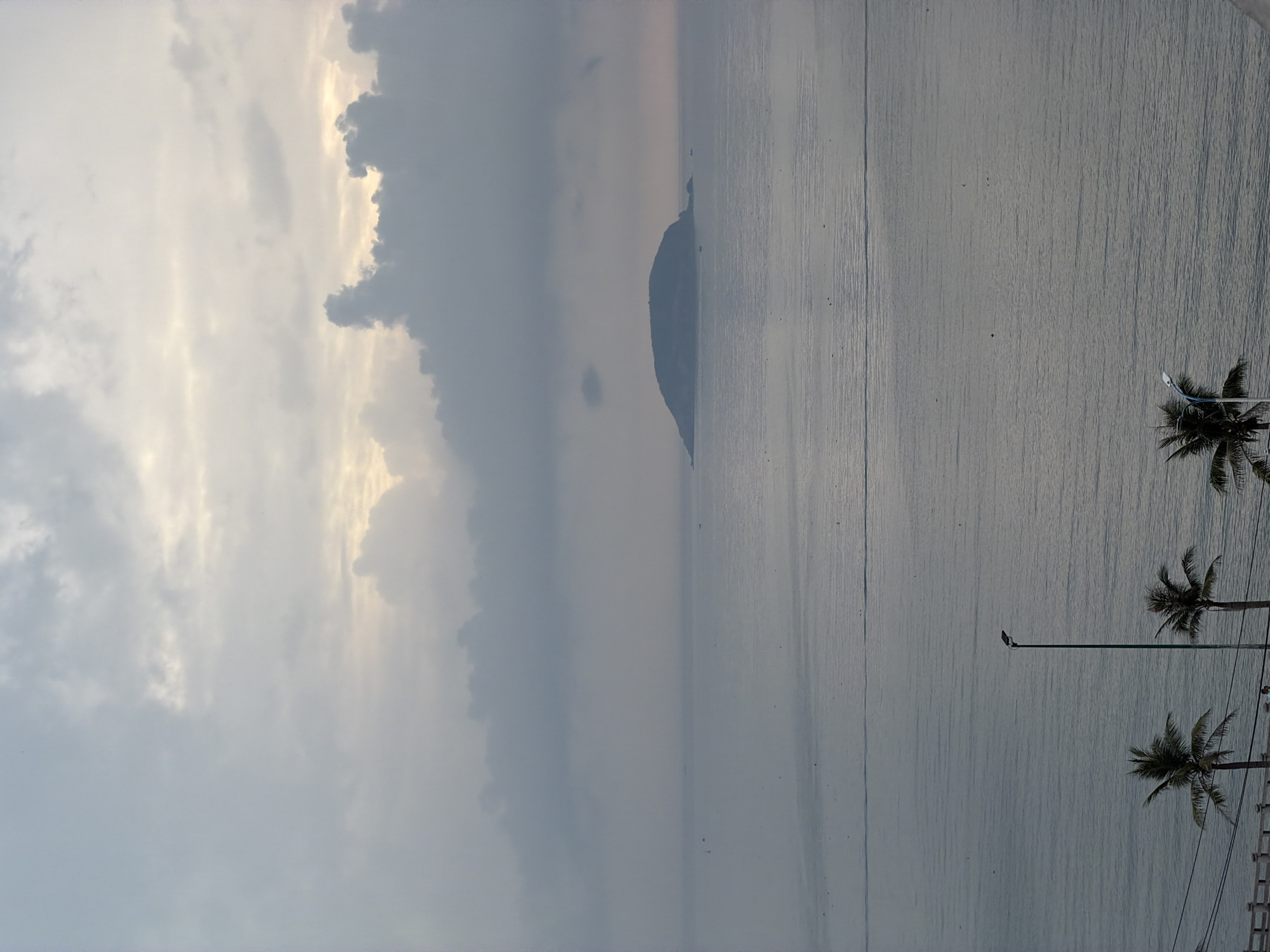 The image depicts a coastal scene in Bac Nha Trang Ward, Vietnam. A body of water, likely the sea, occupies a significant portion of the frame, with a muted grey-blue color. A landmass, potentially an island or peninsula, is visible in the distance. The sky is overcast with heavy, grey clouds, and the scene appears to be at dusk or dawn. On the right, palm trees are visible, along with a street light and some architectural elements.