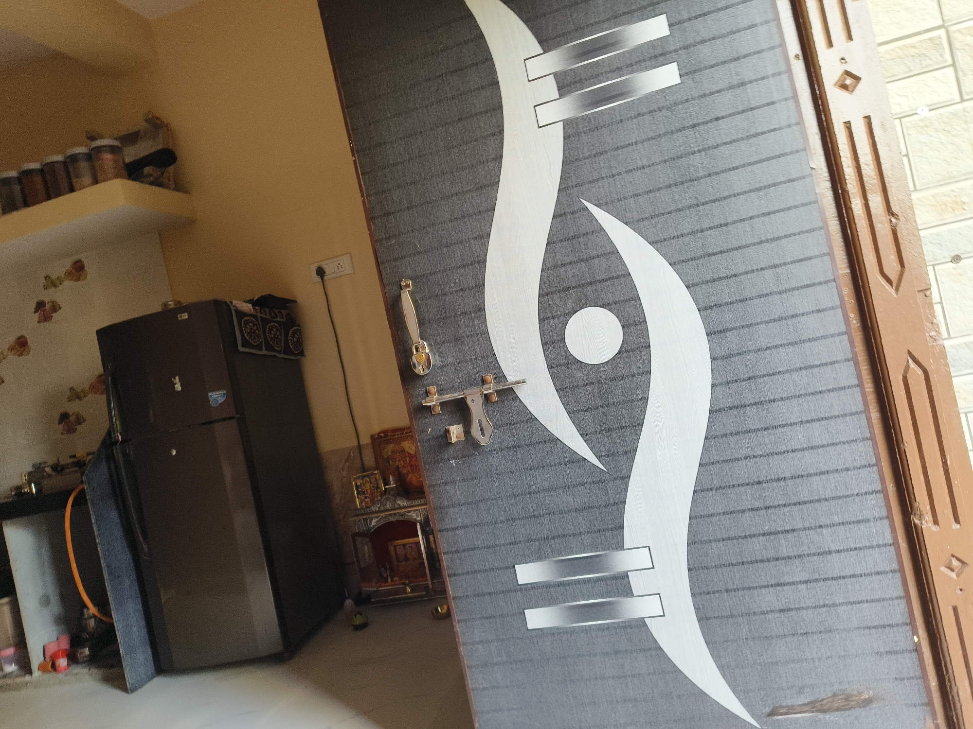 An indoor, slightly angled shot captures a modern door with a grey and white abstract pattern dominating the right side of the frame. The door features a metallic handle and latch. To the left, a domestic interior is visible, likely a kitchen or utility area. A dark-colored double-door refrigerator stands prominently in the center-left. Above it, a pale yellow wall features decorative floral decals, and a wall-mounted shelf holds several jars, likely containing spices or food items. An electrical outlet with a plugged cord is on the adjacent yellow wall. On the floor, near the door, a small, ornate wooden Pooja Mandir (shrine) containing religious figures is present. An orange hose or cable is also on the floor. The setting suggests a home in Dhandhuka, India, under ambient lighting. No individuals are present, and no specific activity is taking place. Text on the refrigerator and jars is illegible.