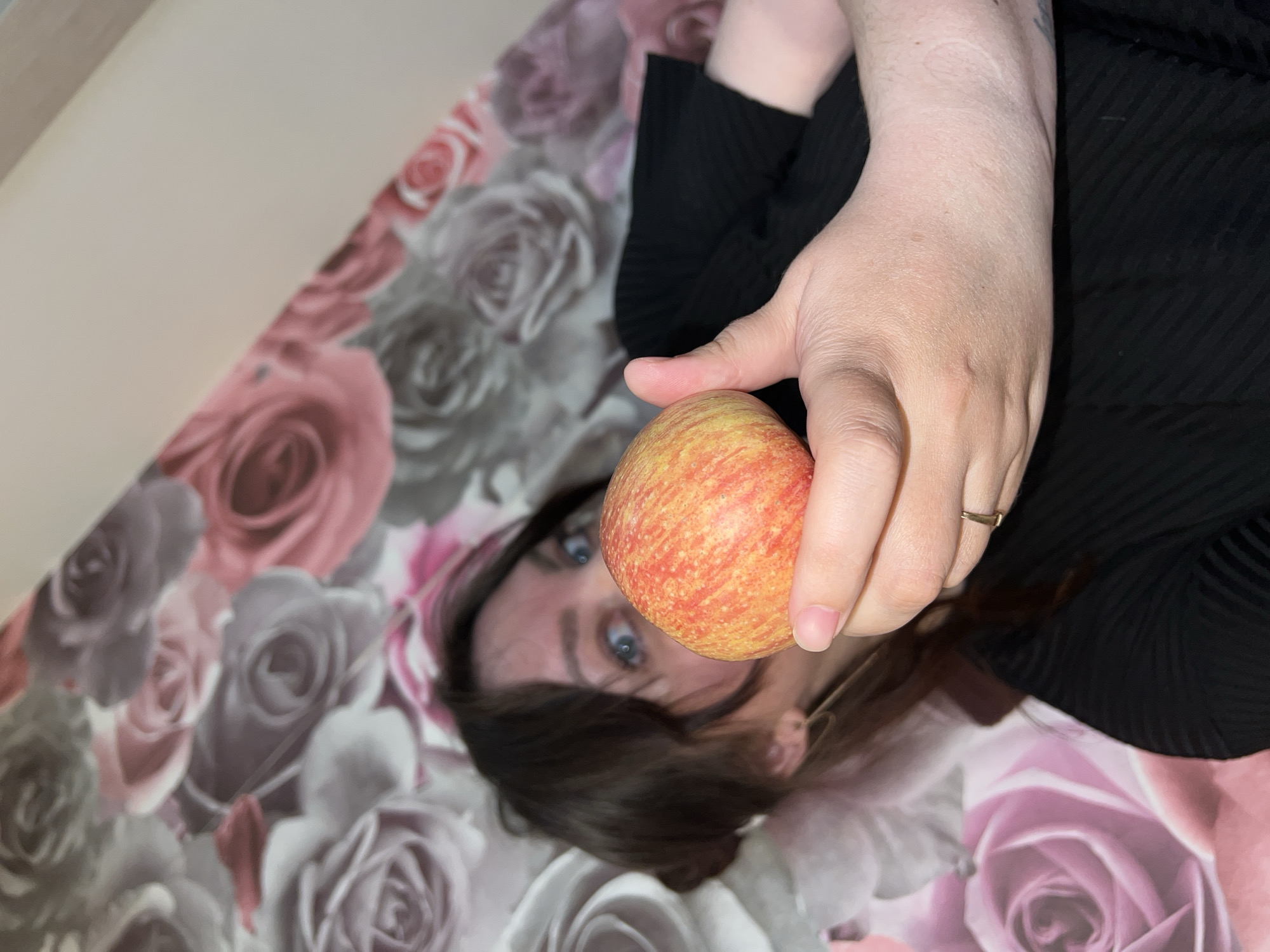 A woman lies on a bed of rose-patterned bedding, holding a red and yellow apple up to her face. She has brown hair and blue eyes, and wears a black top. She has a ring on her right hand. The image is taken from an overhead perspective. The location of Noviy, Russia is not evident in the image.